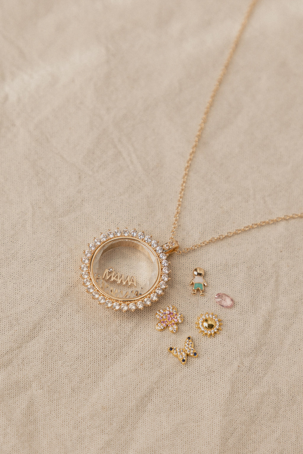 Collar dorado con dije circular decorado con zirconias sobre fondo textil beige, Detalle de collar con colgante redondo brillante acompañado de pequeños charms delicados, Joyería fina en tono dorado: collar con aro de cristales y dijes miniatura a juego, Colgante circular con incrustaciones brillantes y accesorios pequeños en composición elegante, Set de joyería dorada con collar de aro brillante y dijes decorativos sobre superficie neutra.