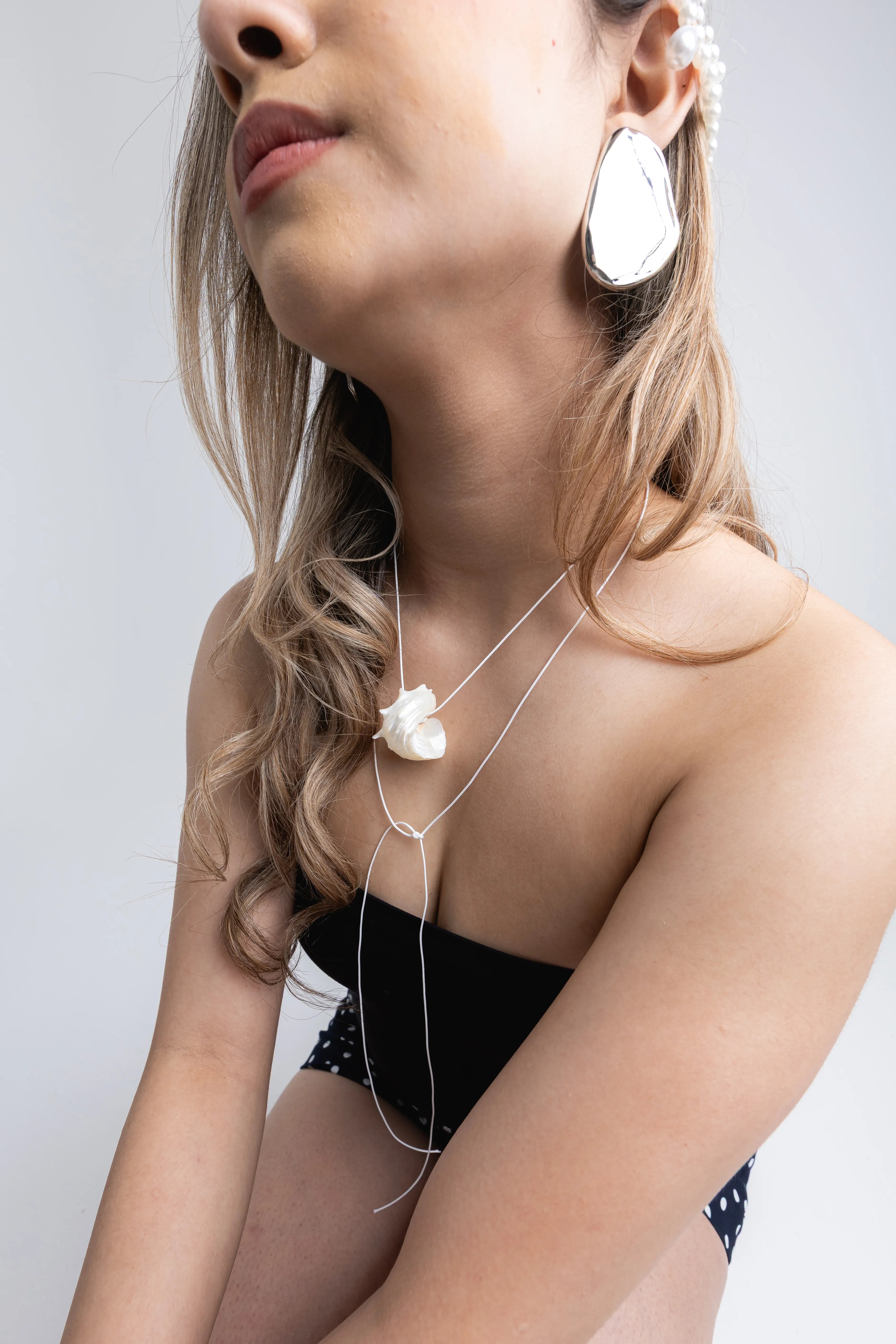 Mujer luciendo un collar de plata con dije de piedra orgánica blanca y aretes grandes plateados, Detalle de joyería artesanal: collar doble de plata, arete ovalado y tocado de perlas en la oreja, Modelo con conjunto de joyas plateadas y conjunto de ropa negra con lunares blancos, Primer plano de collar de plata largo con piedra natural y pendientes de acabado brillante, Look con accesorios de plata: collar de dos capas, aretes XL y detalle de perlas en el cabello.
