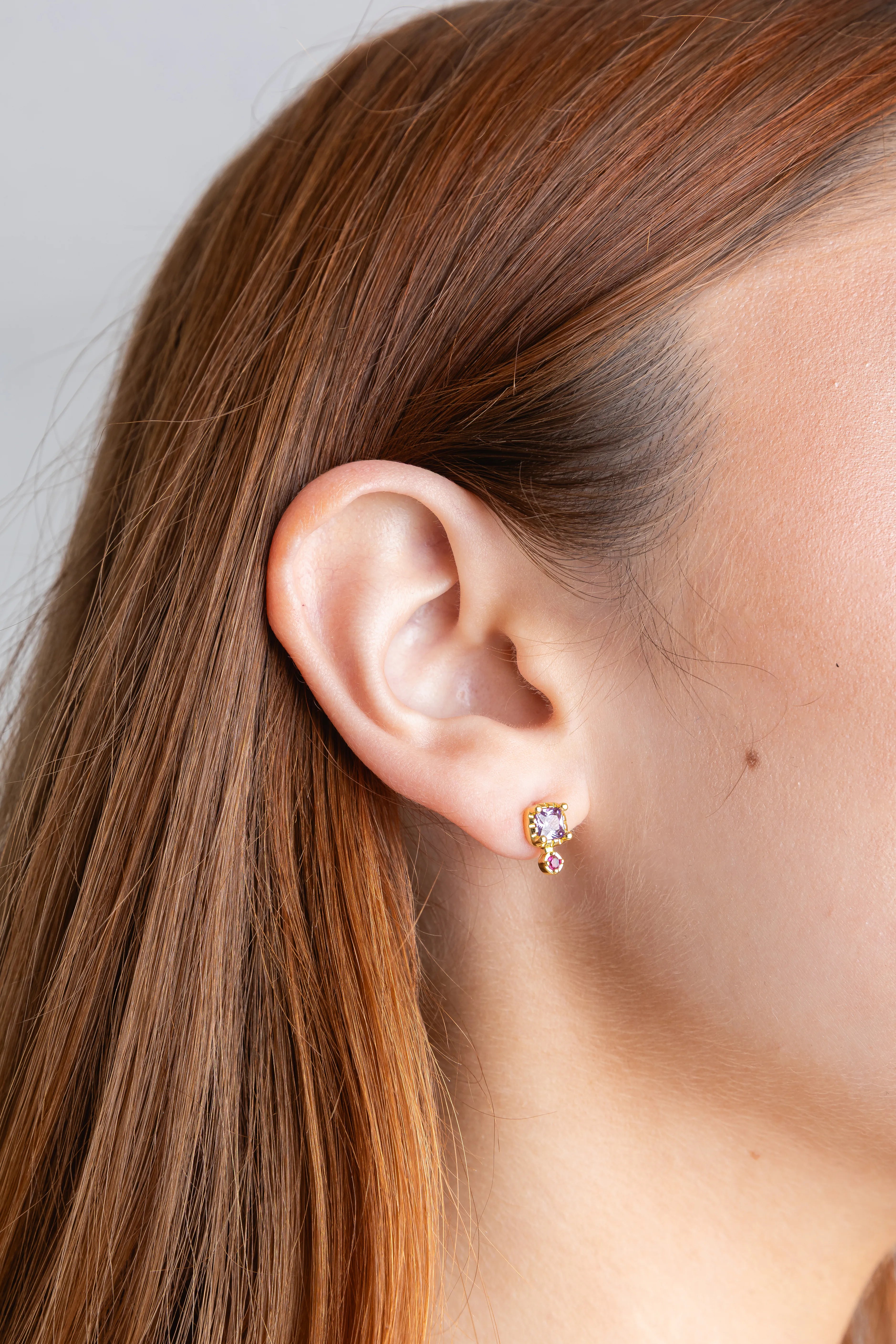 Primer plano de una oreja con un arete pequeño de oro adornado con cristales en color púrpura y rosa, Detalle de joyería delicada: pendiente tipo "stud" dorado con una gema amatista y un brillante inferior, Pendiente minimalista de oro con piedras facetadas en tono violeta sobre el perfil de una modelo, Joyería femenina clásica con un arete de botón dorado y cristales morados resaltando en el lóbulo, Vista de cerca de un accesorio para la oreja con diseño de montura dorada y piedras preciosas lilas.