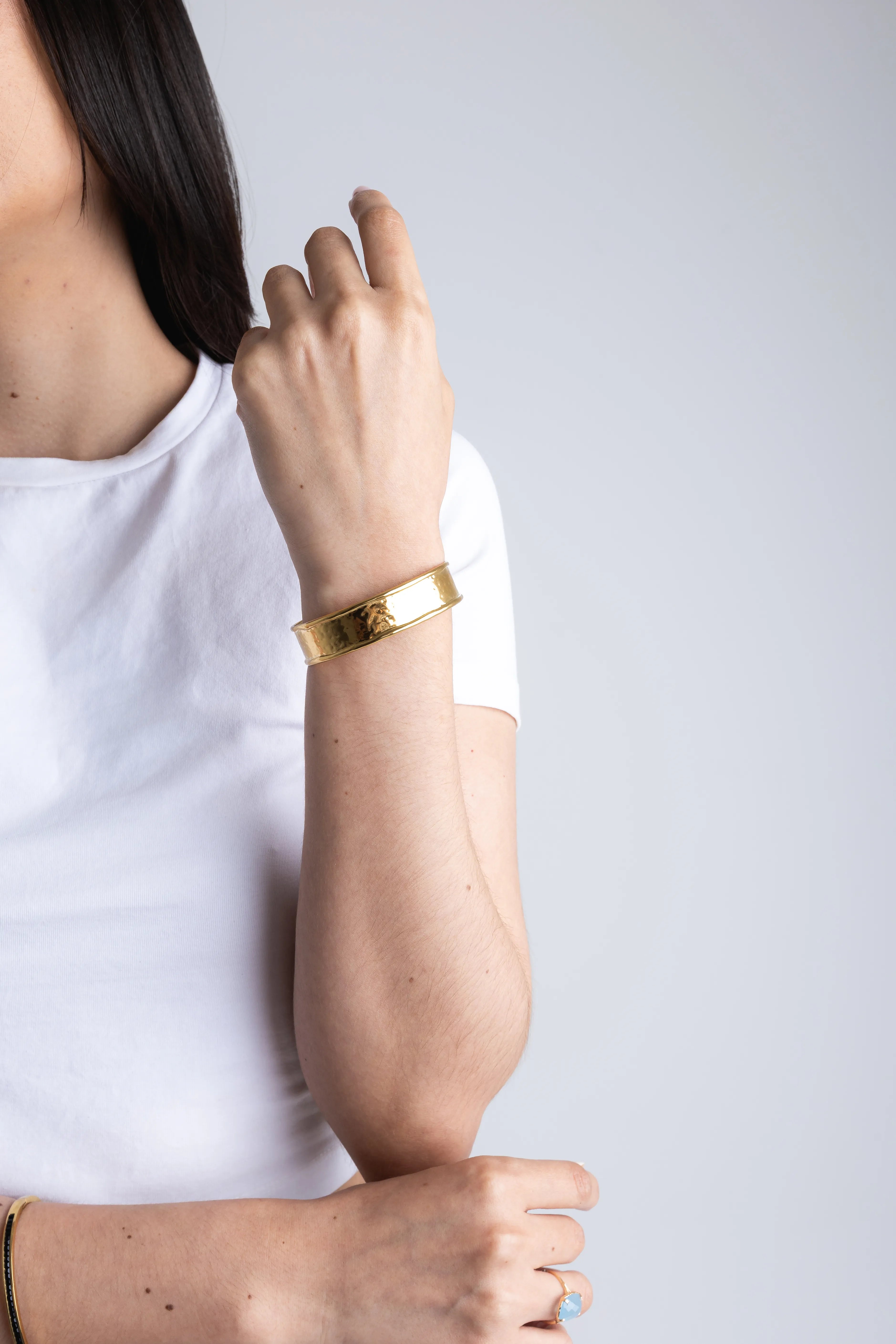 Modelo luciendo un brazalete rígido de oro con acabado martillado y bordes texturizados sobre una camiseta blanca básica, Brazalete ancho de oro de 18k para mujer con diseño artesanal y brillo metálico en primer plano, Detalle de joyería de lujo: pulsera tipo cuff dorada en la muñeca de una mujer, ideal para un look casual elegante, Pulsera ancha dorada con textura grabada y acabado brillante en set de fotografía minimalista, Joyas de oro para mujer: brazalete metálico plano con detalles en los bordes