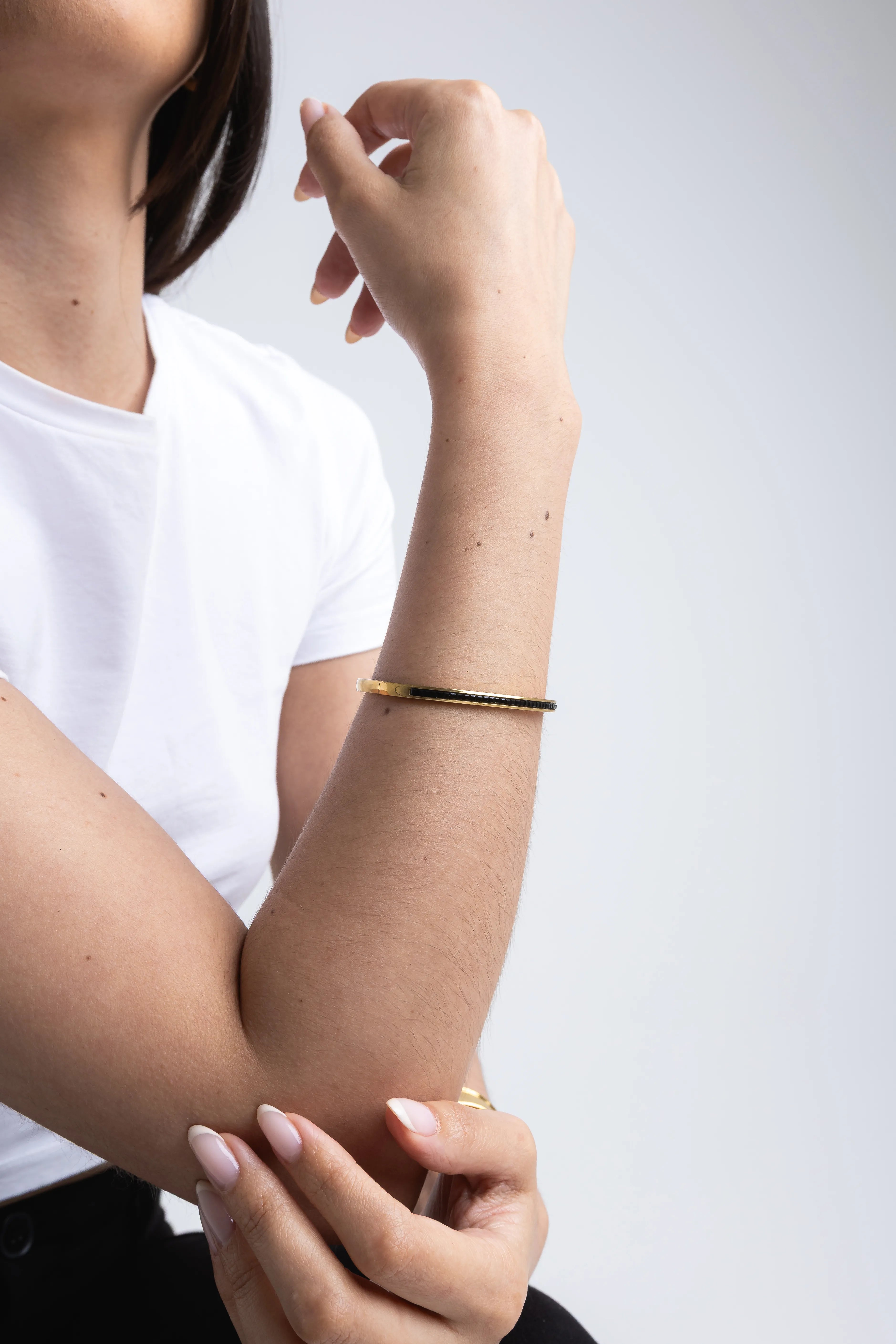 Brazalete delgado de oro con detalle lineal en negro en la muñeca, Pulsera rígida fina dorada con esmalte negro sobre la piel de la modelo, Joyería minimalista: brazalete de oro estrecho con diseño bitono elegante, Detalle de pulsera tipo cuff dorada y negra en conjunto de accesorios, Primer plano de brazalete metálico fino con acabado pulido y línea central negra.