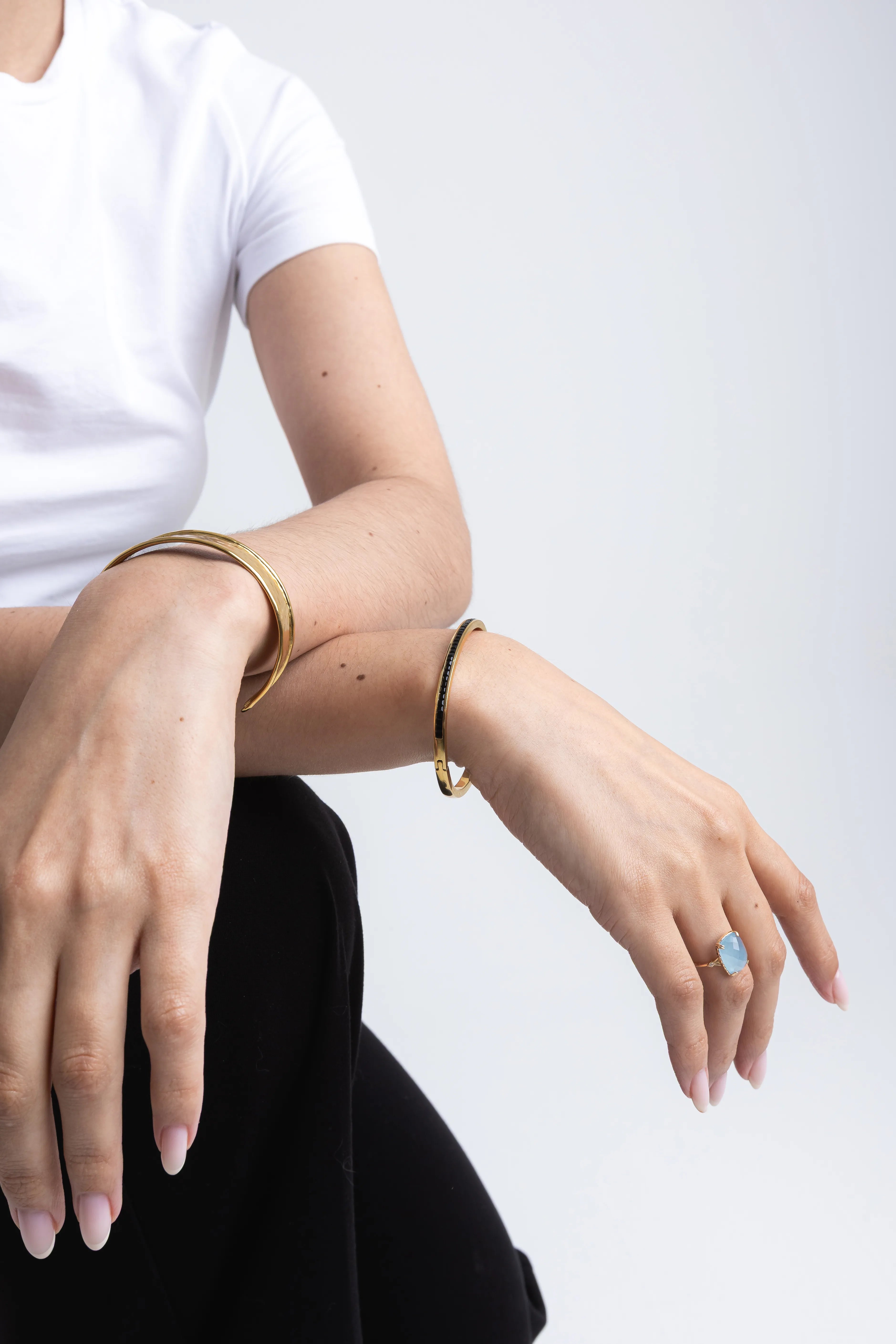 Modelo luciendo un anillo dorado con piedra azul celeste y brazaletes rígidos de oro, Detalle de joyería elegante: anillo con gema azul y pulseras doradas minimalistas, Conjunto de accesorios para mujer con anillo de piedra color aguamarina y brazaletes, Manos de modelo con anillo solitario azul y pulseras de oro sobre fondo blanco, Look de joyería fina con anillo de gema rectangular azul y brazaletes dorados delgados.