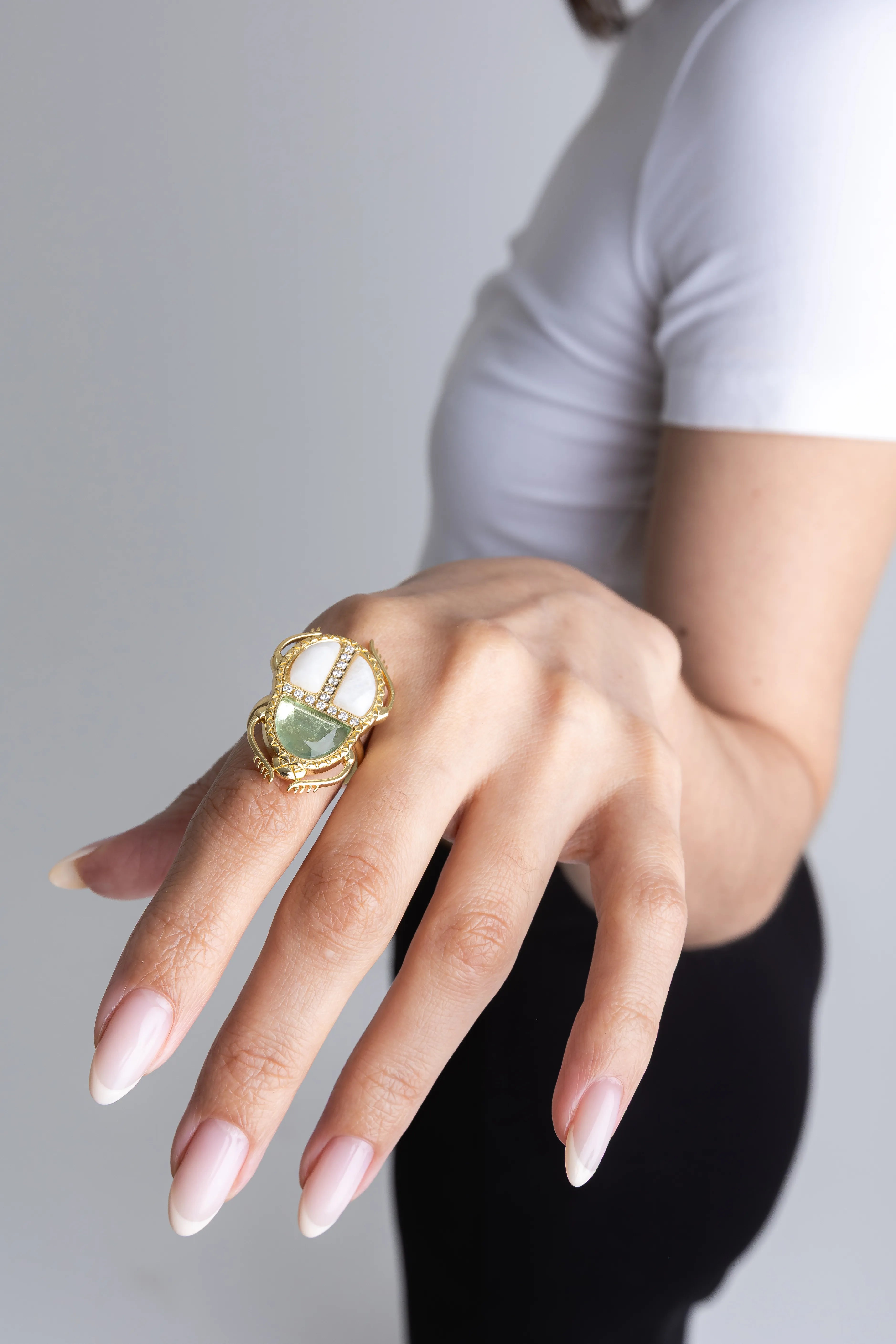 Mano de una mujer con manicura francesa luciendo un anillo dorado grande con forma de escarabajo, Detalle de anillo artesanal dorado con incrustaciones de nácar blanco y una gema verde clara centra, Primer plano de una mano extendida con un anillo llamativo de diseño orgánico en oro y piedras, Anillo de escarabajo detallado con detalles blancos y verdes sobre la mano de una modelo, Accesorios de joyería: anillo XL dorado con diseño de insecto y piedras preciosas sobre fondo neutro.