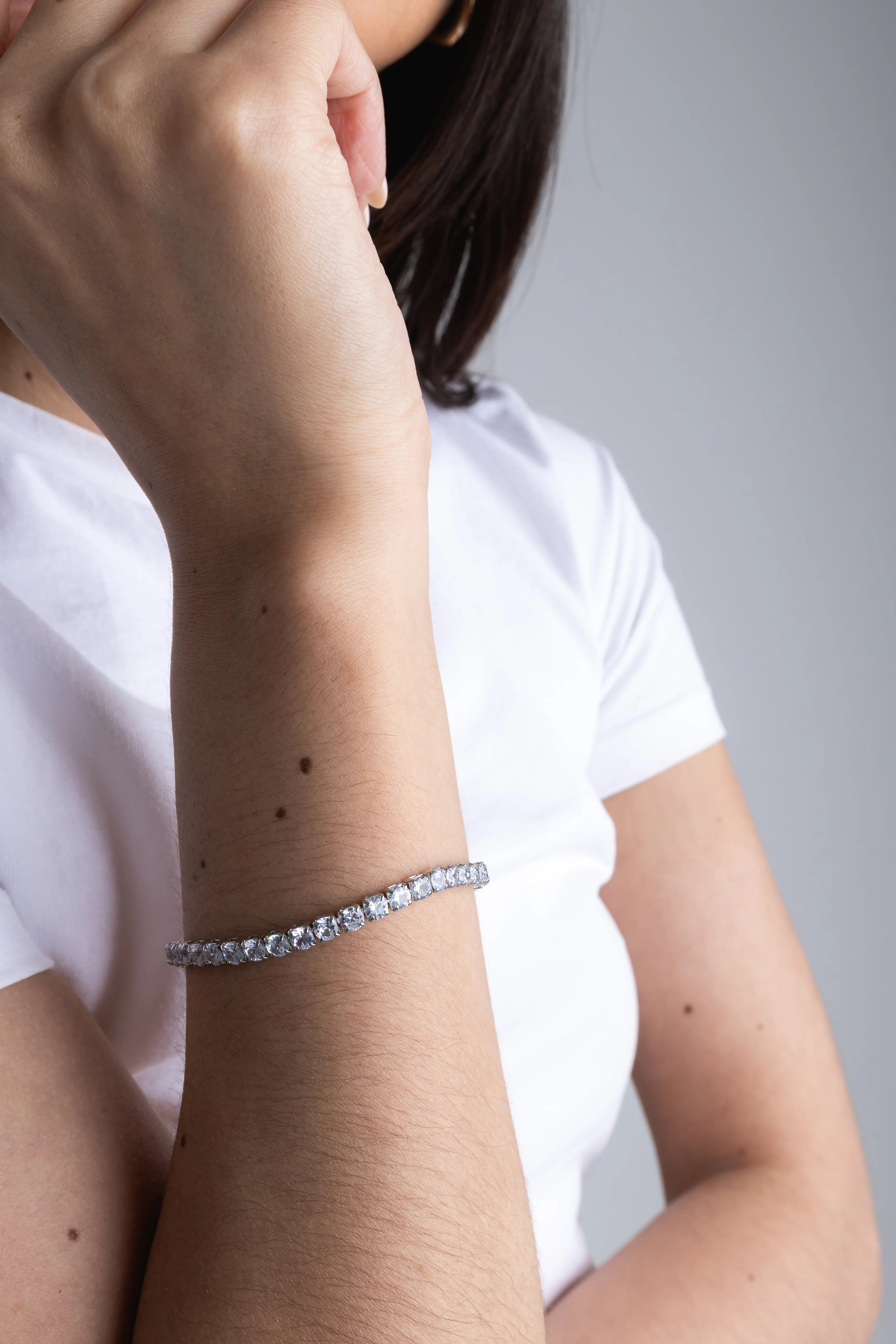 Primer plano de una pulsera de tenis de plata con una fila de cristales brillantes, lucida en la muñeca de una modelo con camiseta blanca, Detalle de joyería clásica: pulsera de eslabones de plata con circonitas cúbicas redondas de corte brillante sobre la piel, 
Mano con manicura natural extendida sutilmente, destacando una pulsera de brillantes de diseño atemporal y elegante, Vista de cerca de un accesorio de muñeca: pulsera riviere de plata esterlina con piedras preciosas transparentes facetadas.