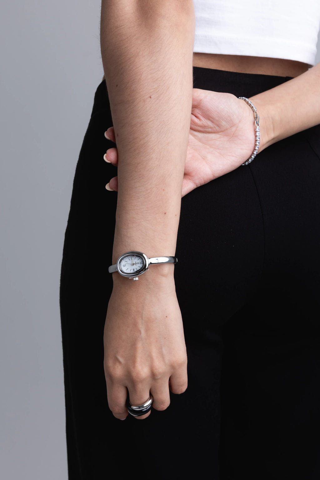 Reloj de pulsera plateado con esfera ovalada pequeña y diseño minimalista en la muñeca, Reloj elegante de acero inoxidable con correa delgada y anillo negro a juego, Detalle de accesorios plateados: reloj vintage ovalado y pulsera de cadena fina en segundo plano, Reloj de dama con acabado en plata y esfera blanca sobre outfit casual de color negro, Primer plano de reloj de mano clásico y discreto con diseño de joyería fina.