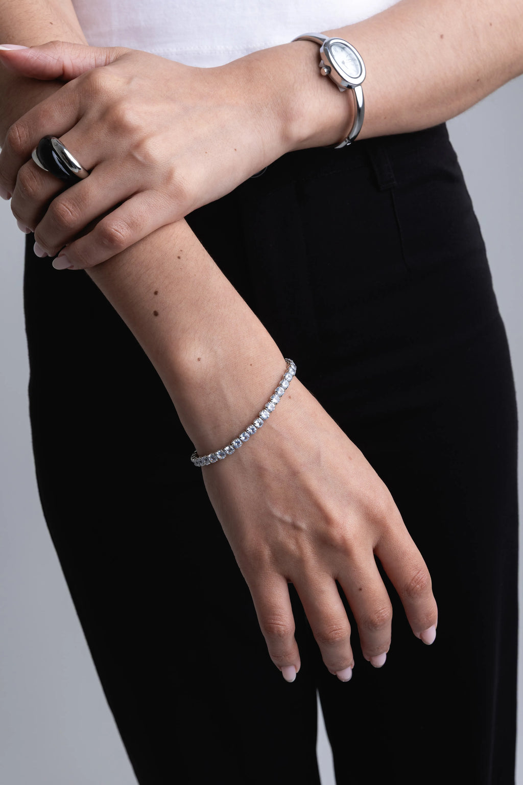 Pulsera tennis de plata con piedras brillantes en la muñeca de una modelo, Anillo grueso entrelazado en color plata y negro sobre el dedo índice, Conjunto de joyería plateada con pulsera de circonitas y anillo moderno, Detalle de pulsera fina con cristales y anillo statement bicolor, Mano de mujer luciendo pulsera brillante de plata y anillo de diseño.