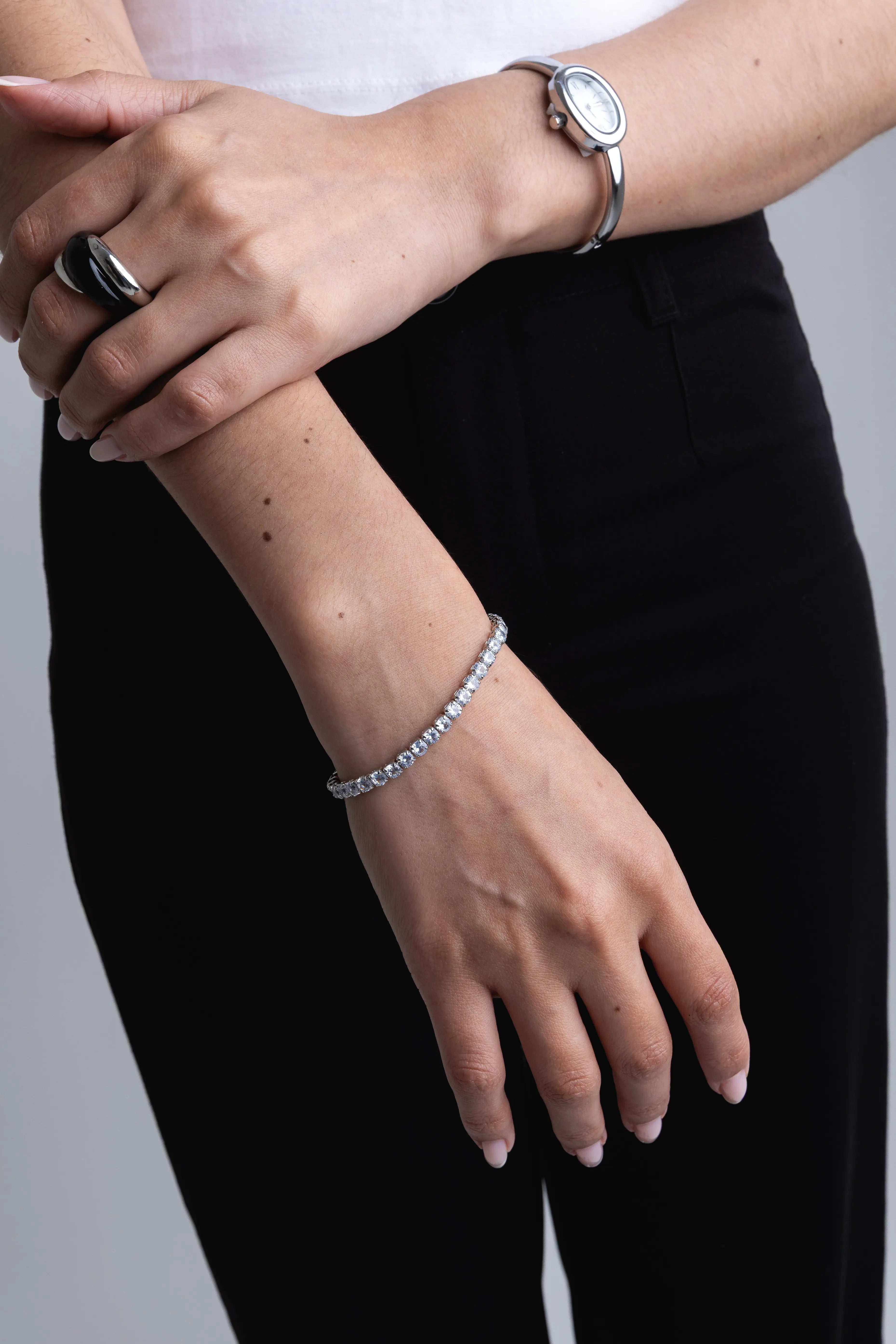 Pulsera tennis de plata con piedras brillantes en la muñeca de una modelo, Anillo grueso entrelazado en color plata y negro sobre el dedo índice, Conjunto de joyería plateada con pulsera de circonitas y anillo moderno, Detalle de pulsera fina con cristales y anillo statement bicolor, Mano de mujer luciendo pulsera brillante de plata y anillo de diseño.