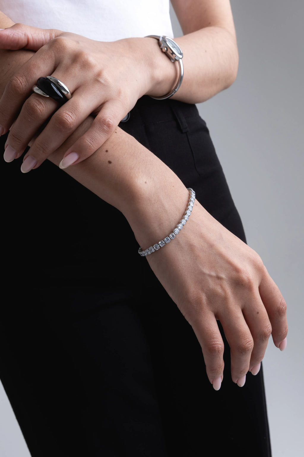 Pulsera tennis de plata con circonitas brillantes de corte marquise en la muñeca de una modelo, Anillo grueso de diseño entrelazado en plata y resina negra en el dedo índice, Conjunto de joyería de plata para mujer: pulsera de piedras brillantes y anillo moderno bicolor, Detalle de pulsera delicada con cristales transparentes y anillo de plata texturizada con negro, Look de joyería contemporánea con pulsera de brillo fino y anillo Statement en plata y negro.