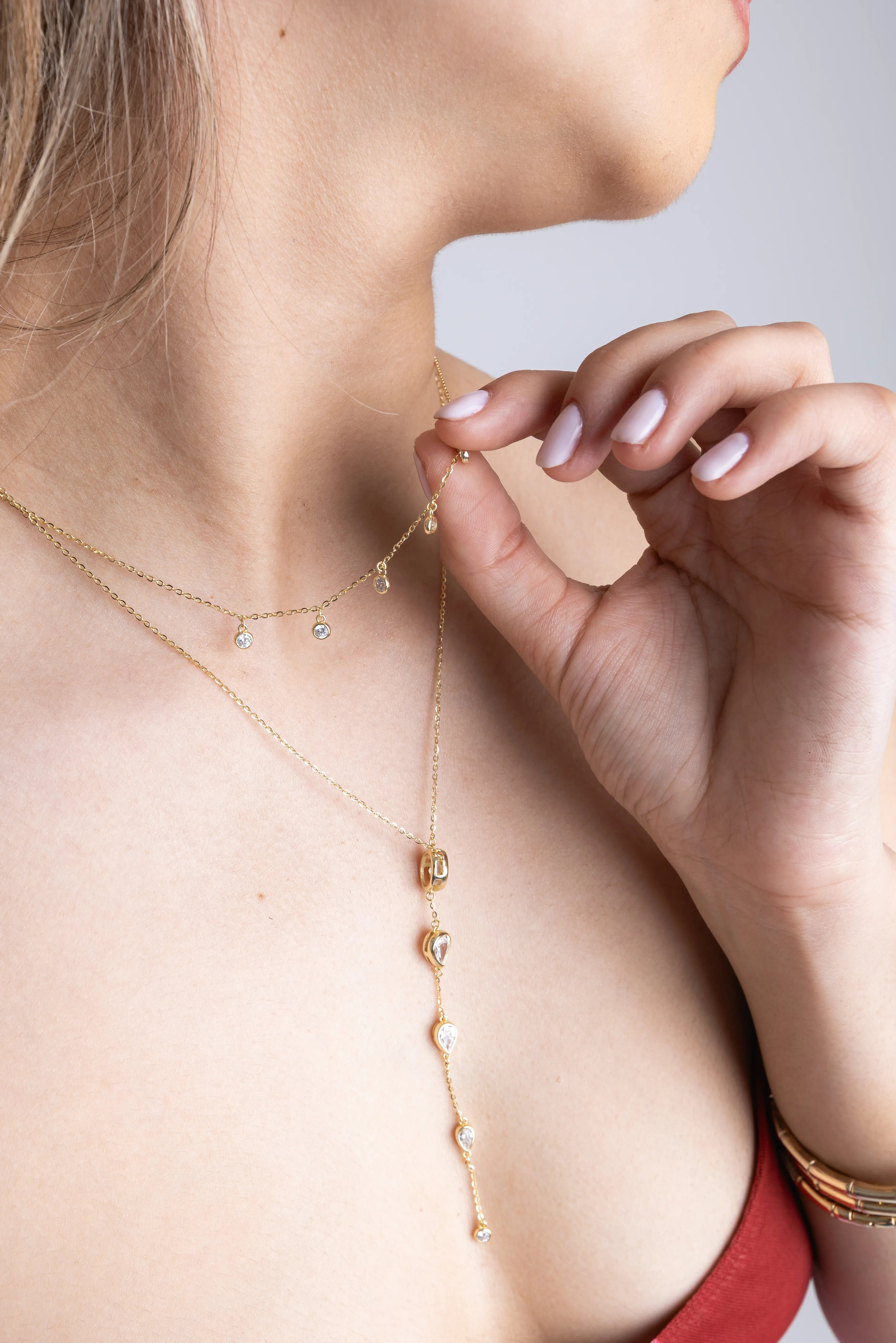 Mujer ajustando collar dorado en capas con dijes brillantes sobre escote, Detalle de collares finos en tono dorado con pequeños cristales y colgante alargado, Joyería delicada en capas: cadenas doradas con dijes sutiles y diseño elegante, Primer plano de collar largo con elementos brillantes y cadenas finas combinadas, Accesorios de joyería femenina con collares en capas y colgantes minimalistas dorados.
