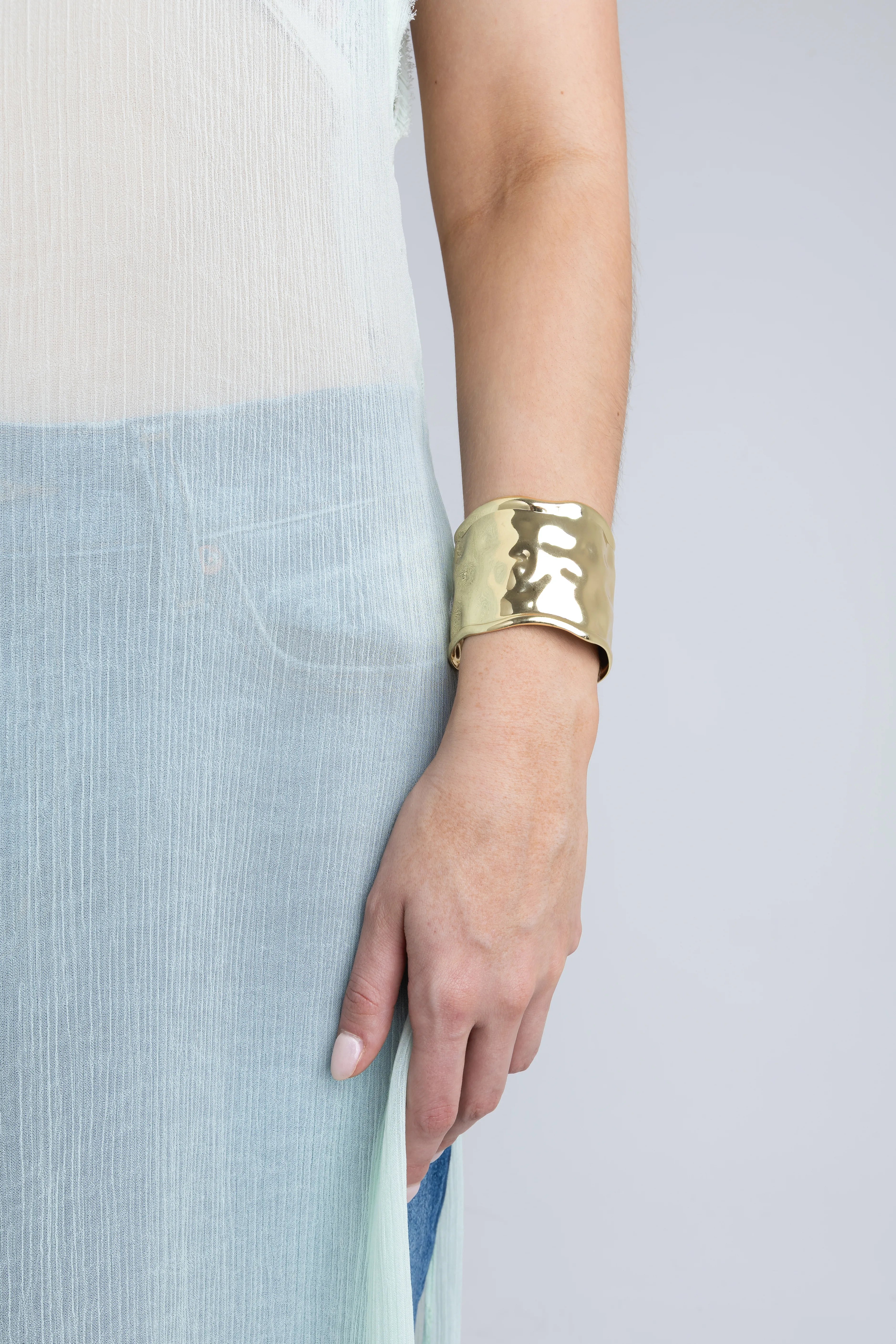 Brazalete ancho de oro con acabado martillado y textura irregular en la muñeca de una modelo, Pulsera tipo puño dorada con efecto de metal orgánico y brillo satinado, Accesorio de joyería: brazalete grueso de oro con bordes ondulados sobre fondo minimalista, Detalle de brazalete rígido dorado estilo contemporáneo complementando un look casual, Pulsera ancha dorada con superficie texturizada que refleja la luz de forma natural.