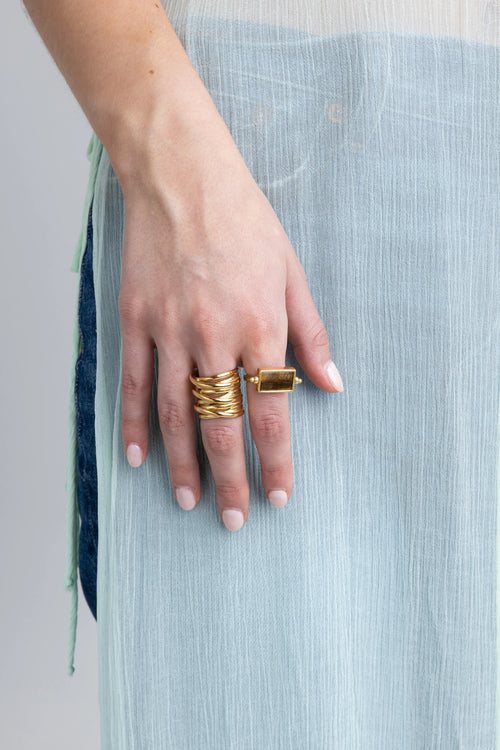 Mano de mujer con anillo ancho de oro martillado y anillo de piedra rectangular color ámbar, Combinación de anillos dorados: uno grueso con diseño de bandas y otro con gema café translúcida, Detalle de joyería en la mano: anillos de oro con texturas orgánicas y piedras naturales, Anillo robusto dorado estilo artesanal junto a un anillo con gema rectangular sobre fondo azul claro, Set de anillos de oro para mujer con acabados rústicos y piedras cuadradas elegantes.