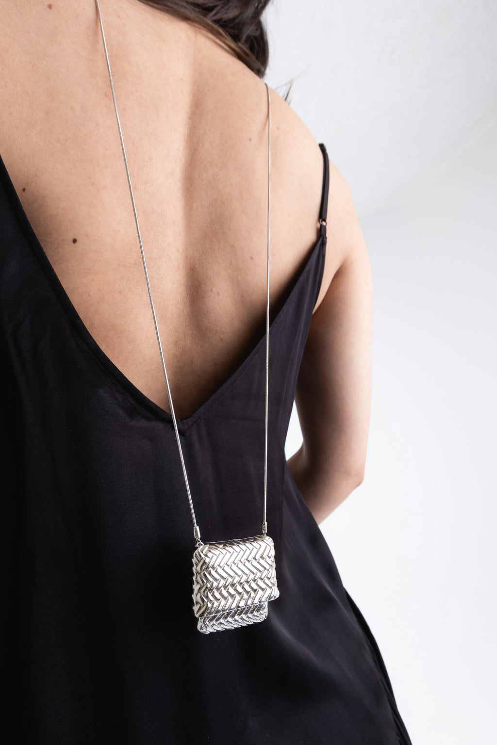 Collar largo de plata con dije rectangular de diseño tejido cayendo sobre la espalda, Cadena delgada plateada con colgante estilo bolso miniatura texturizado en acabado brillante, Joyería para espalda: collar largo con dije de plata labrada sobre vestido negro satinado, Detalle de accesorio plateado con colgante geométrico y patrón de espiga en relieve, Primer plano de collar de plata largo y minimalista con dije cuadrado artesanal.