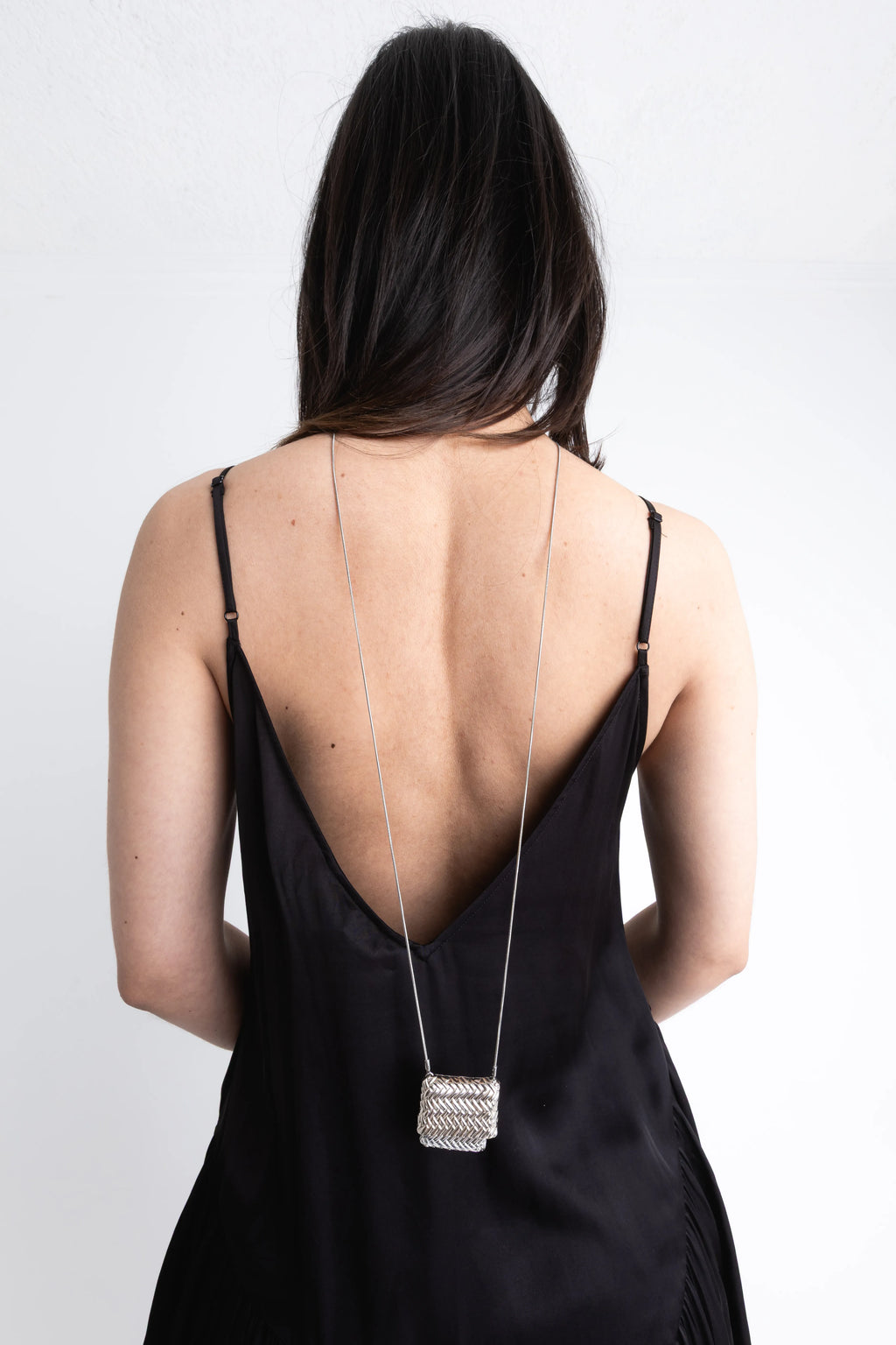 Vista trasera de modelo con un collar largo de plata que cae sobre un escote profundo en la espalda, Collar de espalda con dije cuadrado plateado y textura de malla metálica brillante, Accesorio de joyería para vestidos con escote posterior: cadena fina y colgante rectangular de plata, Collar largo "back-drop" con un pequeño dije tejido que resalta sobre un vestido negro, Diseño de joyería minimalista: cadena plateada larga con adorno geométrico central para la espalda.