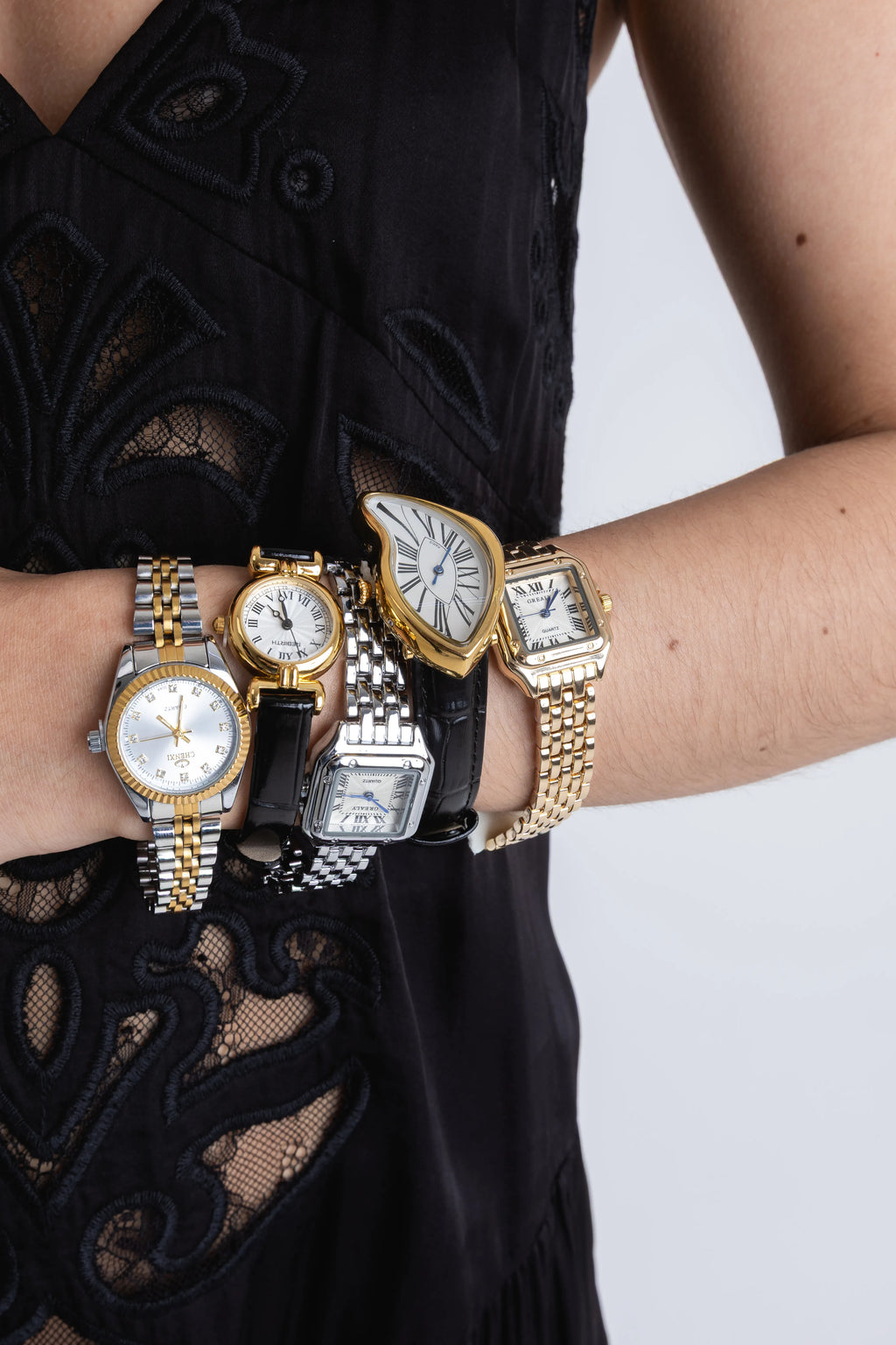 Brazo de una mujer luciendo una colección de cinco relojes de pulsera diferentes con acabados en oro y plata, Composición de relojes de lujo: modelos con extensibles metálicos bicolor, correas de cuero negro y esferas de formas irregulares, Detalle de relojes para dama, incluyendo un diseño de esfera derretida estilo surrealista y relojes clásicos cuadrados y redondos, Varios relojes de pulsera elegantes apilados en el brazo de una modelo vistiendo un conjunto de encaje negro.