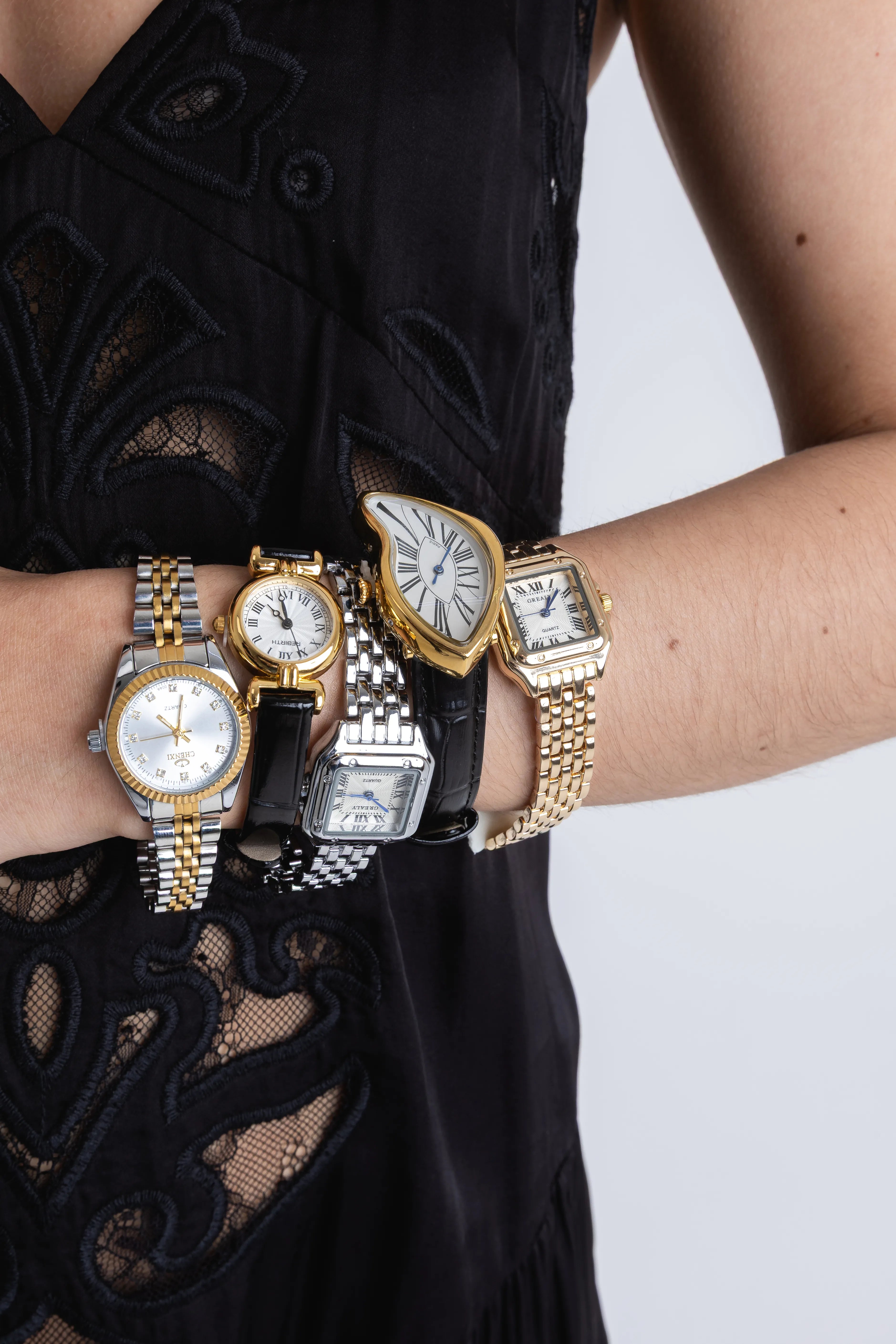 Brazo de una mujer luciendo una colección de cinco relojes de pulsera diferentes con acabados en oro y plata, Composición de relojes de lujo: modelos con extensibles metálicos bicolor, correas de cuero negro y esferas de formas irregulares, Detalle de relojes para dama, incluyendo un diseño de esfera derretida estilo surrealista y relojes clásicos cuadrados y redondos, Varios relojes de pulsera elegantes apilados en el brazo de una modelo vistiendo un conjunto de encaje negro.