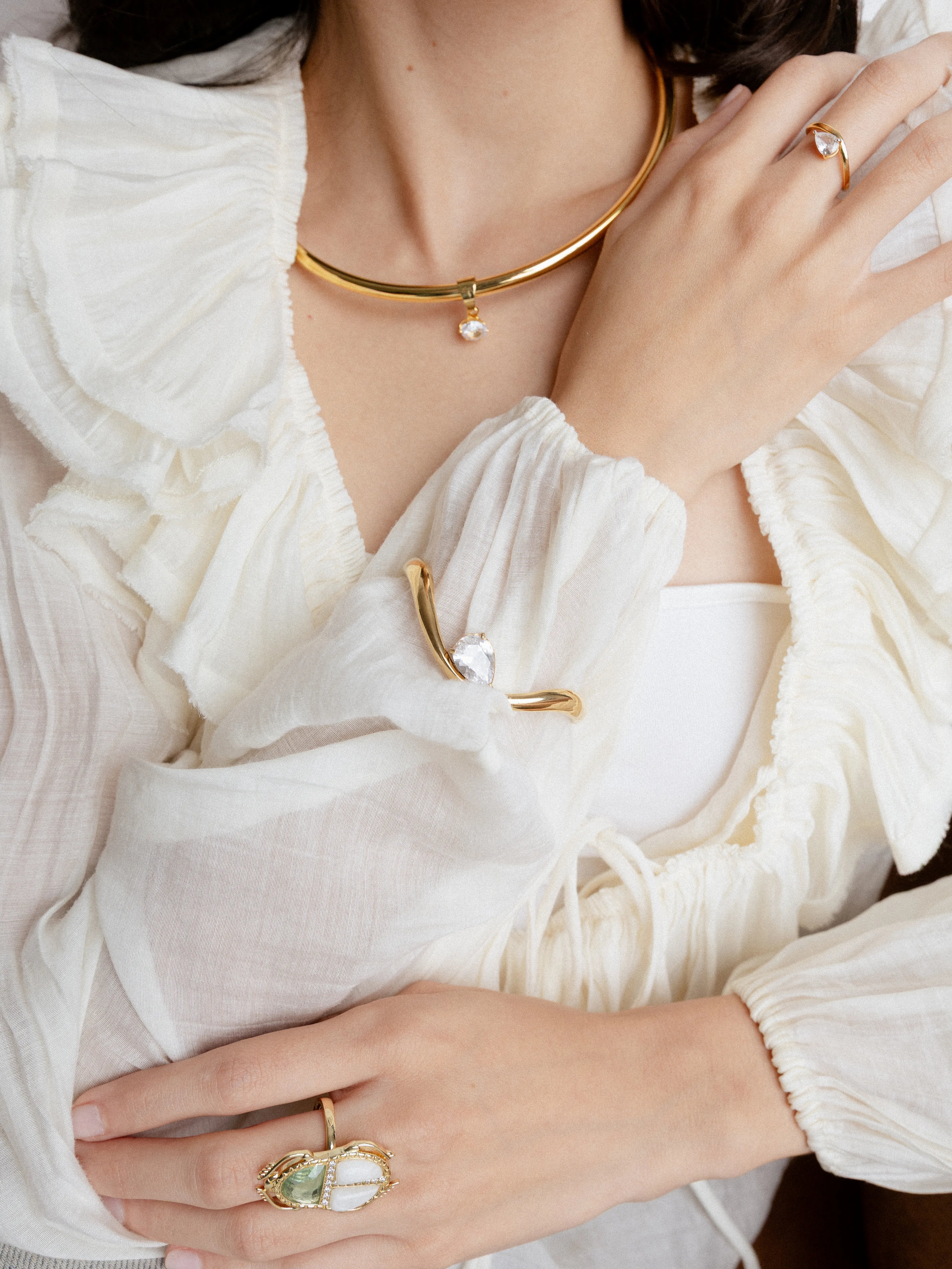 Gargantilla rígida de oro con un pequeño dije de cristal circular en el centro, Mujer luciendo un set de joyería dorada con piedras claras, incluyendo collar rígido y anillos, Detalle de collar tipo "torque" dorado y anillo con gema en forma de gota sobre blusa blanca, Joyería minimalista en oro: gargantilla fina y sortijas con brillantes transparentes, Primer plano de accesorios dorados con piedras naturales y diseño contemporáneo.