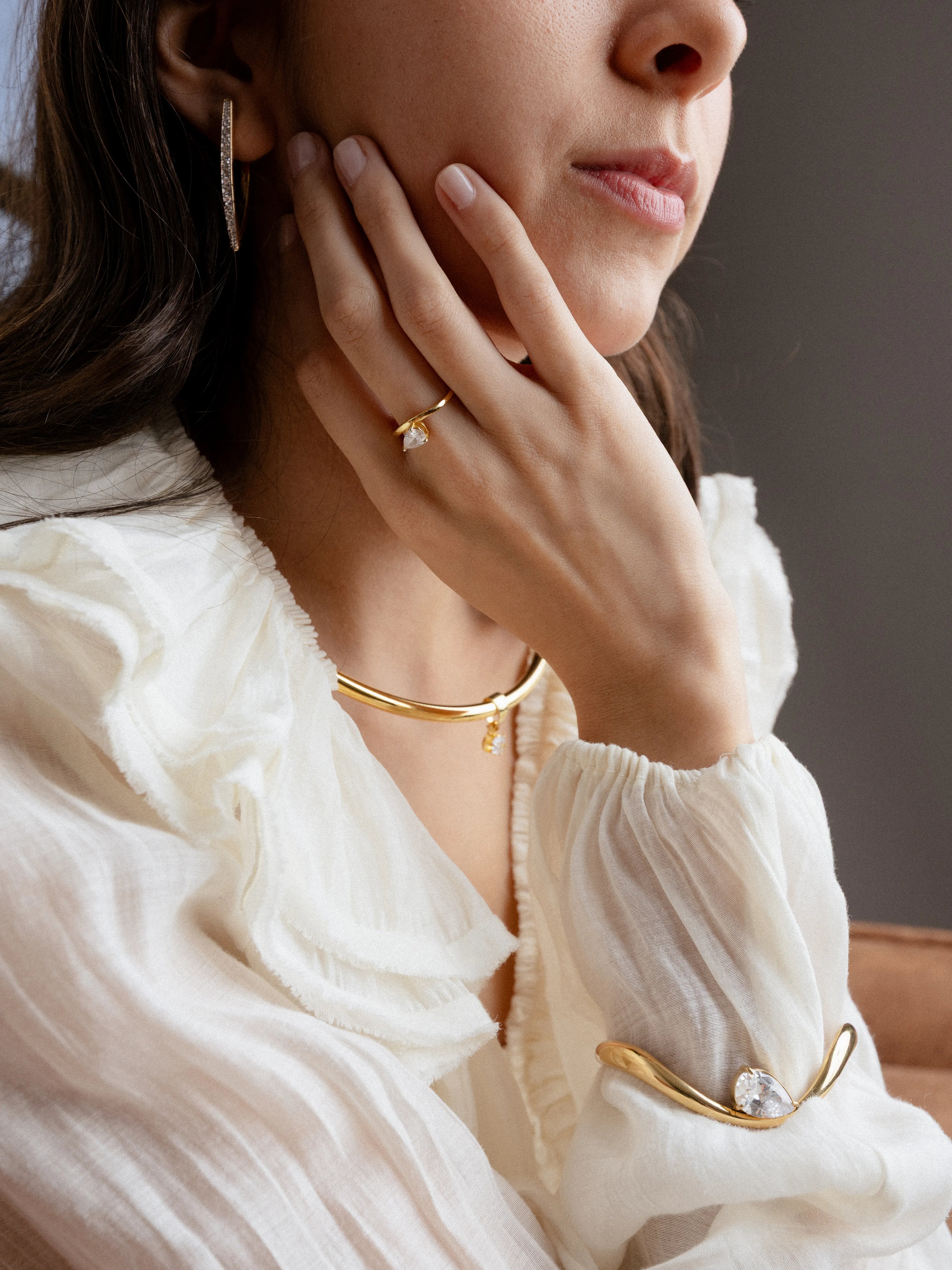 Mujer posando con un conjunto de joyería dorada que incluye una gargantilla rígida, anillos con cristales y un brazalete abierto, Gargantilla fina de oro con dije pequeño y brillante sobre una blusa blanca de textura ligera, Detalle de accesorios de joyería: anillo con gema en forma de gota y brazalete dorado con cristal central, Set de joyería elegante en oro amarillo y piedras transparentes lucido por una modelo, Primer plano de pendientes largos de oro y gargantilla minimalista complementando un look.