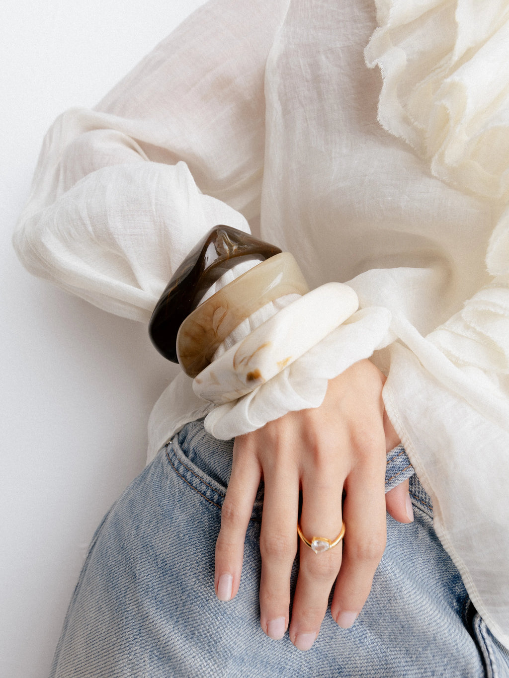 Mano de mujer con un conjunto de tres brazaletes anchos en tonos café, beige y blanco veteado, Pulseras gruesas de resina o piedra natural combinadas con un anillo de oro y gema transparente, Detalle de accesorios estilo boho: brazaletes rígidos de gran tamaño y anillo delicado sobre jeans, Set de joyería de diseño orgánico con pulseras tipo "bangle" y anillo con piedra en forma de gota, Primer plano de brazaletes artesanales en colores neutros y sortija dorada minimalista.