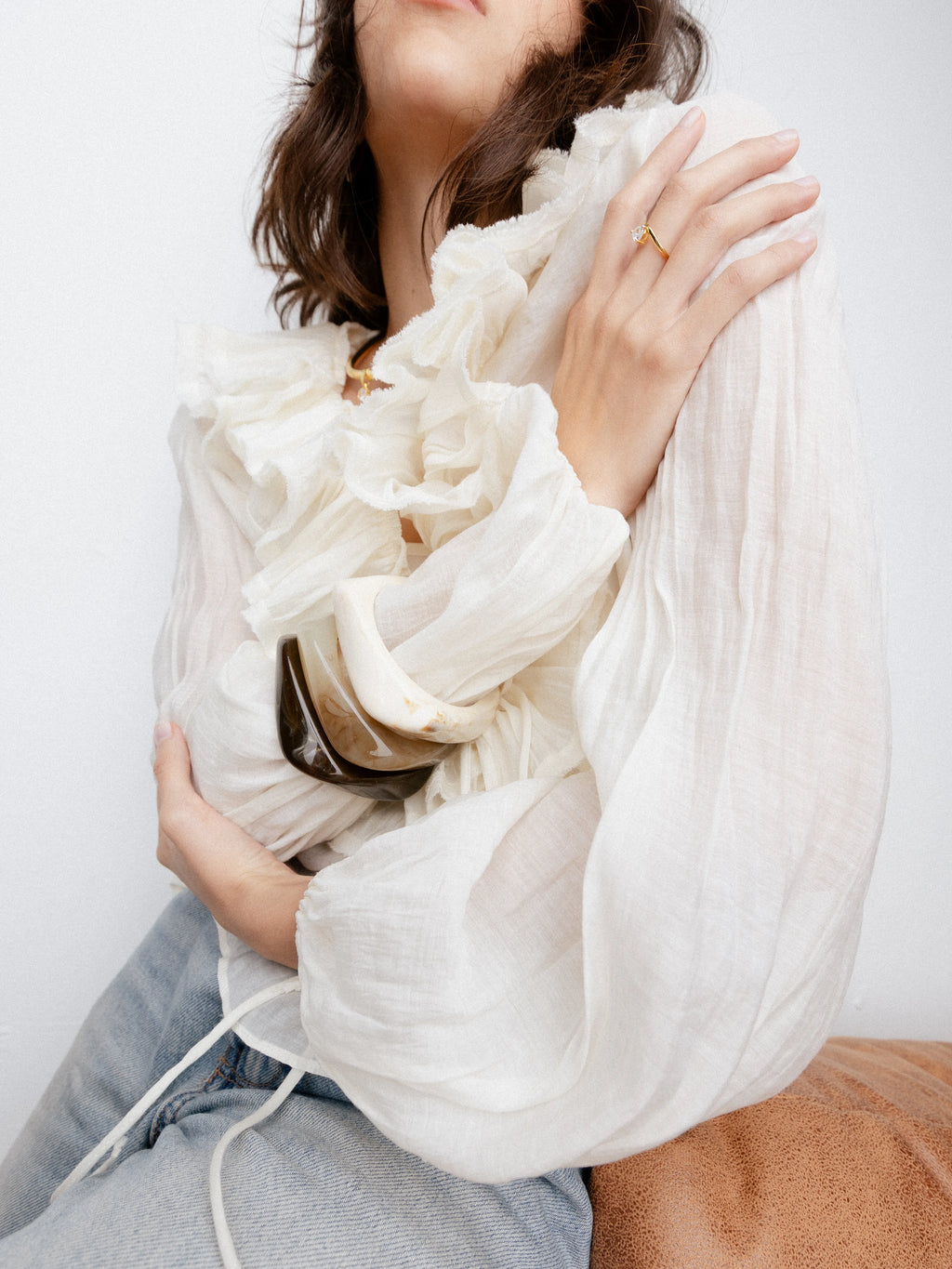 Mujer posando con blusa de volantes blanca, luciendo brazaletes de resina anchos en tonos tierra y un anillo dorado fino, Detalle de accesorios bohemios: pulseras rígidas en colores café y crema junto a un anillo con piedra pequeña, Set de brazaletes de diseño orgánico y sortija de oro en una modelo con estilo romántico y casual, Brazaletes tipo "bangle" de gran tamaño en tonos neutros complementando un look de textura ligera, Primer plano de joyería artesanal: pulseras gruesas veteadas y anillo minimal
