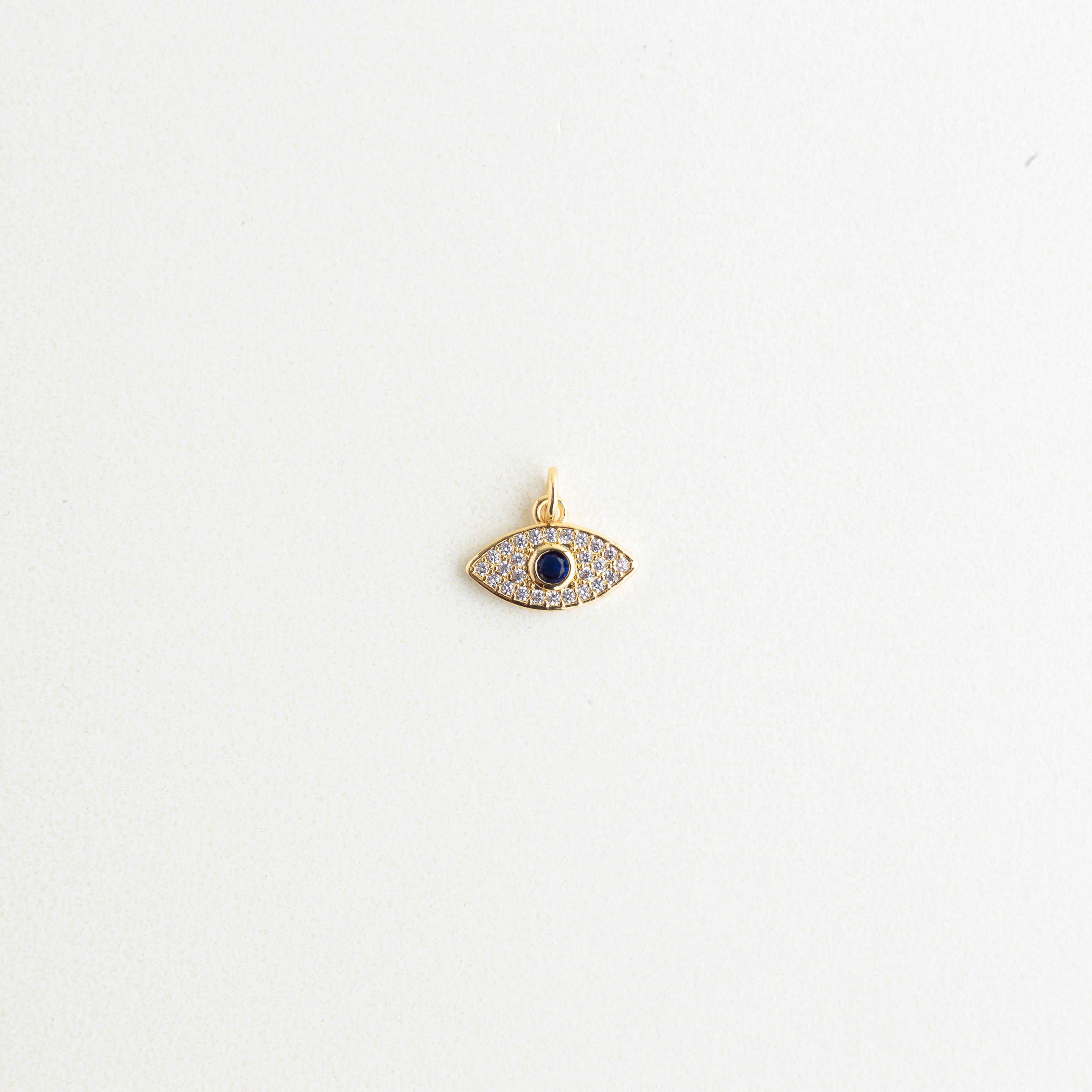Dije mini de ojo turco dorado con pavé de zirconias transparentes y piedra azul central, Charm de cobre enchapado en oro en forma de ojo místico de Amalaya Jewelry, Dije protector de ojo turco de 1 cm para personalizar cadenas y pulseras de moda, Amuleto de joyería dorado con zirconia azul zafiro sobre fondo blanco texturizado, Charm para armar joyería con silueta de ojo místico de la suerte y cristales.