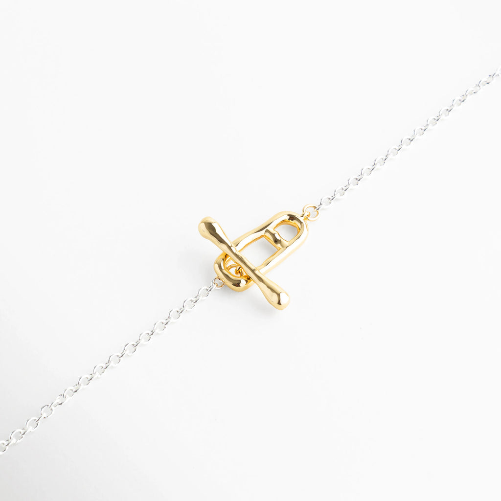 Primer plano de cierre tipo T-Bar enchapado en oro de 18K sobre cadena de plata, Detalle de broche marinero frontal dorado con acabado brillante en collar bi-tono, Cierre de barra 'T' dorado de 18K integrado en cadena de eslabones finos platinados, Zoom a la pieza central dorada de un collar minimalista con base de cobre y oro, Detalle de joyería moderna mostrando el contraste entre el cierre dorado y la cadena blanca.