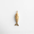 Charm dorado en forma de pez articulado con textura detallada y ojo con toque de color, diseño divertido y elegante que simboliza abundancia y buena fortuna, ideal para agregar movimiento y estilo a cualquier look