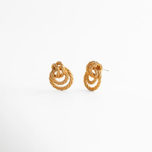 aretes dorados con diseño tridimensional entrelazado, vista lateral de aretes con relieve y textura marcada, aretes compactos con estructura curva y elegante, joyería dorada con diseño escultórico y profundidad, aretes de moda con acabado brillante y forma envolvente