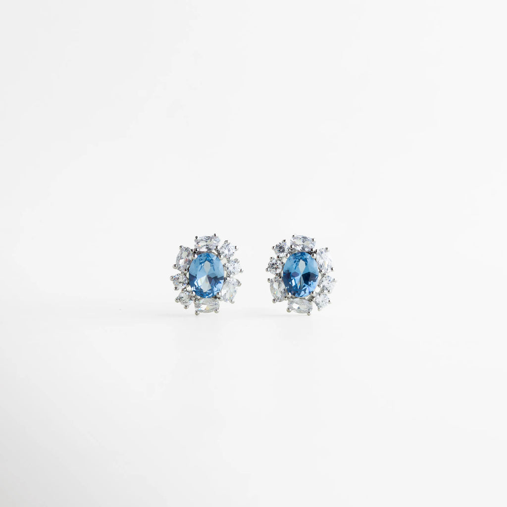 Aretes stud plateados con piedra azul central y halo de zirconias, Aretes pequeños elegantes con piedra azul brillante, Aretes clásicos plateados con zirconias y gema azul, Studs plateados delicados con diseño tipo halo, Aretes finos con piedra azul para uso diario, Aretes minimalistas elegantes con brillo sofisticado