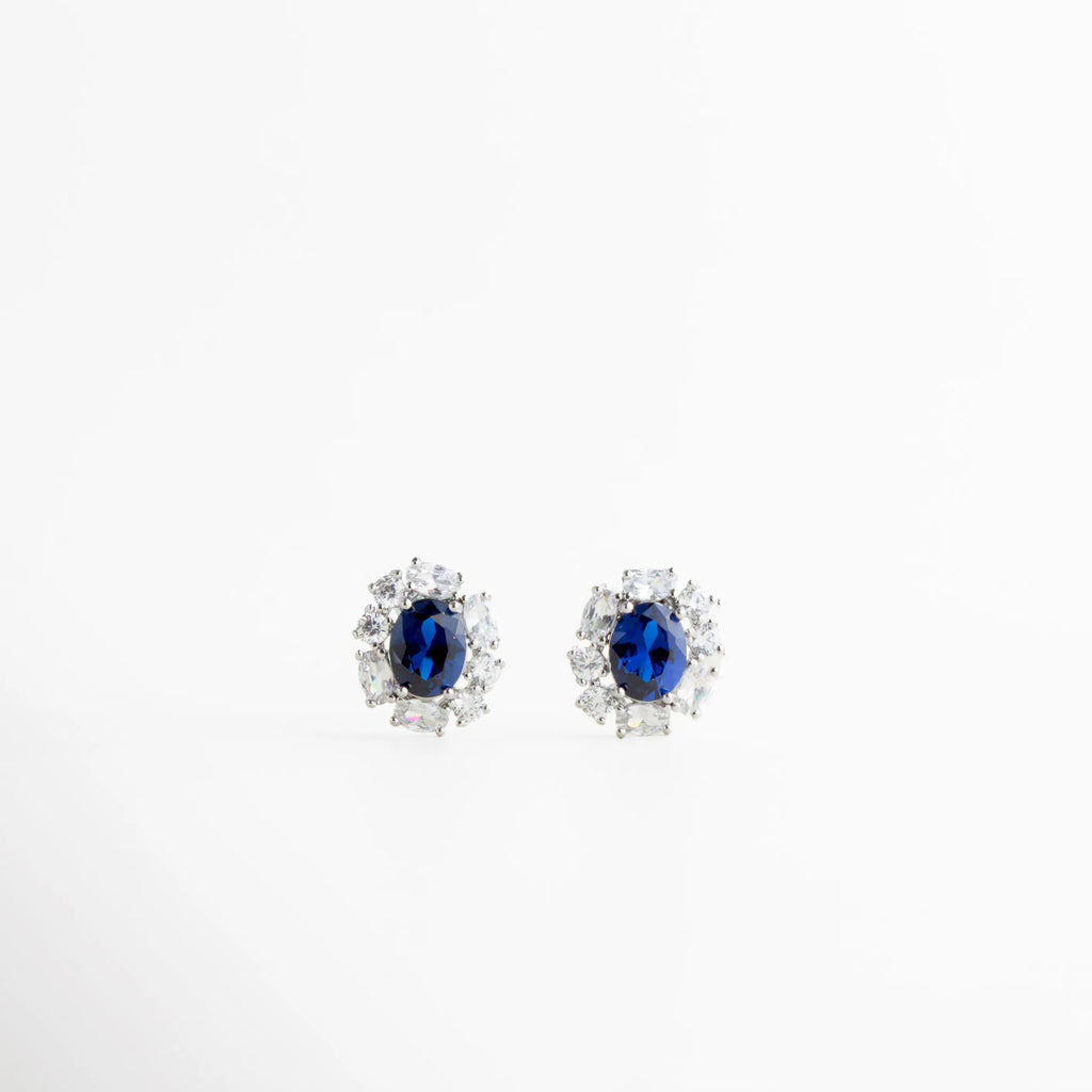 Aretes stud plateados con piedra azul oscuro y halo de zirconias, Aretes elegantes con gema azul profundo, Studs plateados clásicos con piedra azul intensa, Aretes pequeños con zirconias y diseño sofisticado, Aretes finos plateados con brillo profundo, Aretes elegantes con piedra azul oscuro central