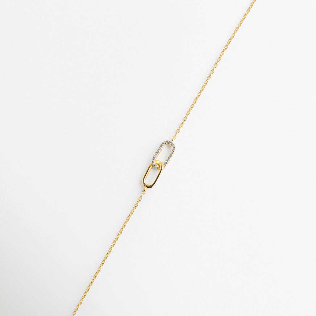 Pulsera de plata 925 con baño de oro y dije de dos eslabones entrelazados, uno con circonias, Detalle de pulsera fina dorada con diseño de eslabón tipo "link" decorado con cristales, Primer plano de pulsera minimalista con incrustaciones de circonia en eslabón central, Joyería de plata ley 925: pulsera dorada con detalle de doble eslabón brillante, Pulsera delicada de cadena fina con dije de unión en oro y circonias transparentes.