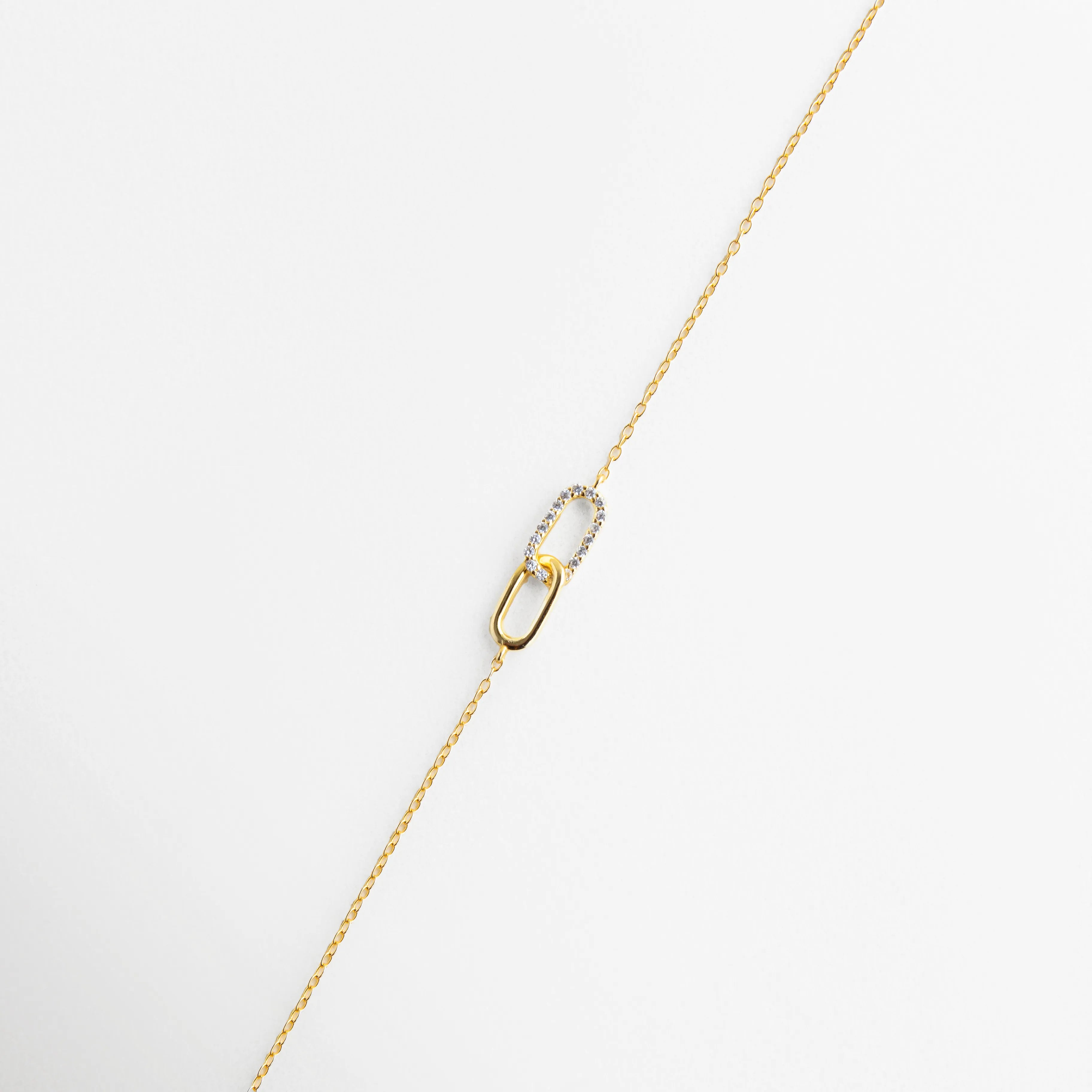Pulsera de plata 925 con baño de oro y dije de dos eslabones entrelazados, uno con circonias, Detalle de pulsera fina dorada con diseño de eslabón tipo "link" decorado con cristales, Primer plano de pulsera minimalista con incrustaciones de circonia en eslabón central, Joyería de plata ley 925: pulsera dorada con detalle de doble eslabón brillante, Pulsera delicada de cadena fina con dije de unión en oro y circonias transparentes.