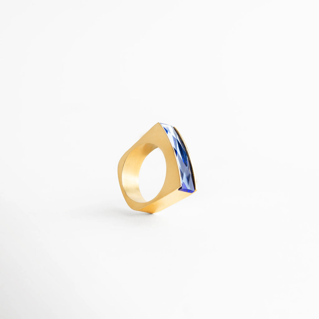 Anillo dorado con piedra azul facetada, diseño geométrico contemporáneo con reflejos intensos, ideal para looks modernos.