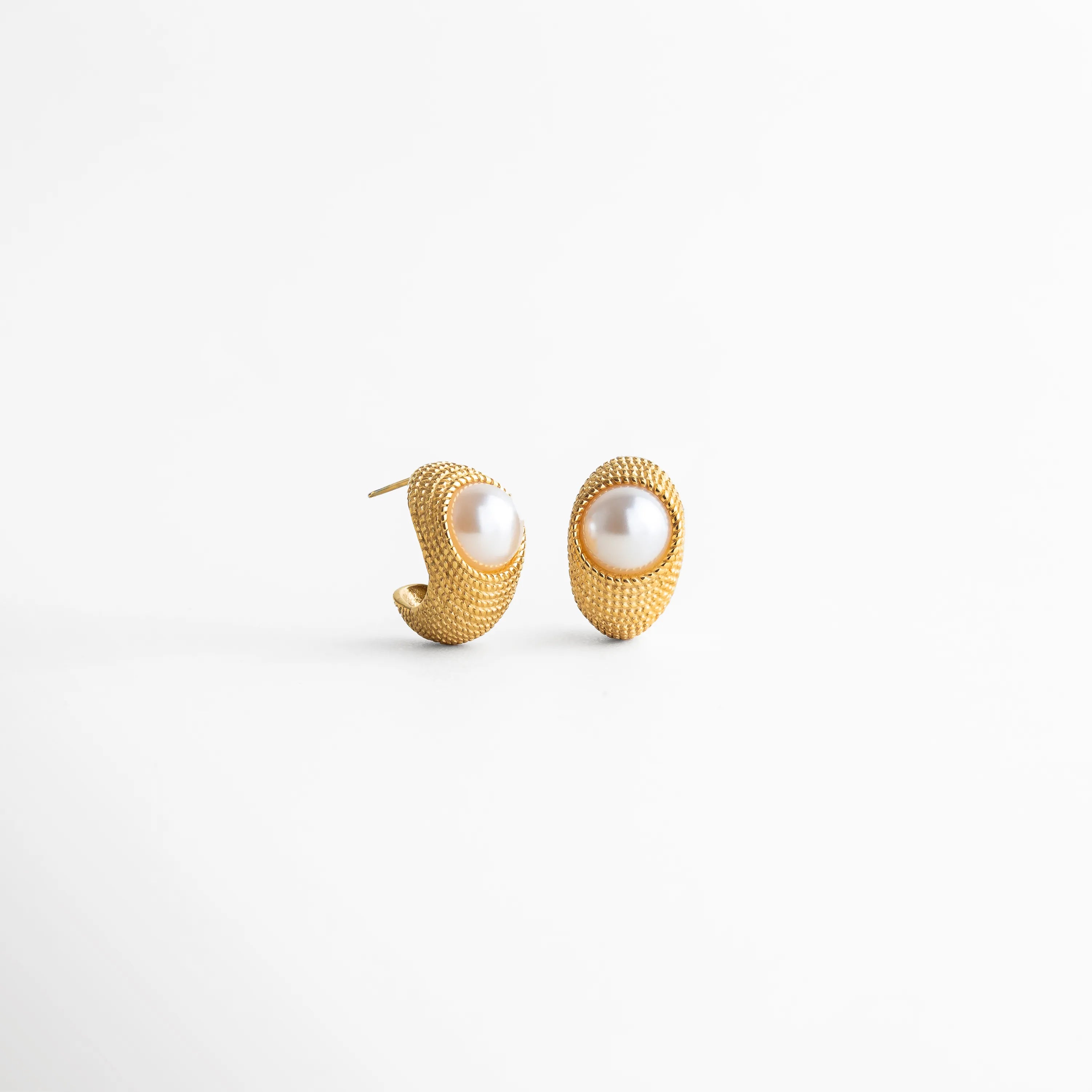 Vista lateral de aretes dorados con perla sintética y textura detallada en acabado oro 18K, Perfil de aretes ovalados en acero inoxidable con chapa dorada y perla incrustada, Aretes Bimba vista lateral mostrando relieve texturizado y perla sintética central, Pendientes dorados con diseño envolvente alrededor de perla, vista de perfil, Detalle lateral de aretes con acabado texturizado y perla sintética 