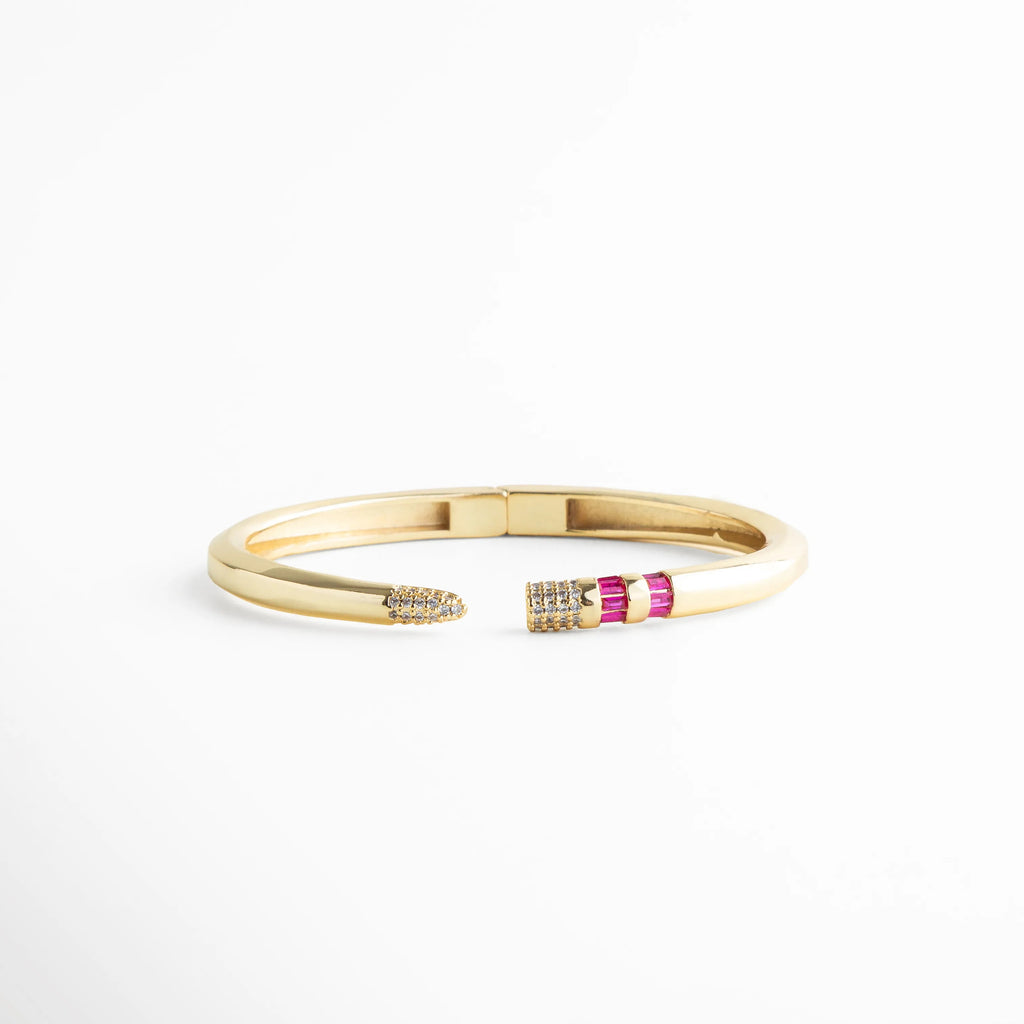 Brazalete rígido dorado con puntas abiertas decoradas con circonias fucsia y blancas, Pulsera de cobre con chapa de oro 18K y detalle de piedras baguette en color magenta, Vista frontal de brazalete abierto con incrustaciones de circonia cúbica y acabado pulido, Joyería elegante: brazalete dorado con diseño de puntas asimétricas y cristales brillantes, Brazalete tipo "cuff" con bisagra central y detalles de pedrería fina en tonos rosa y cristal.