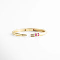 Brazalete rígido dorado con puntas abiertas decoradas con circonias fucsia y blancas, Pulsera de cobre con chapa de oro 18K y detalle de piedras baguette en color magenta, Vista frontal de brazalete abierto con incrustaciones de circonia cúbica y acabado pulido, Joyería elegante: brazalete dorado con diseño de puntas asimétricas y cristales brillantes, Brazalete tipo "cuff" con bisagra central y detalles de pedrería fina en tonos rosa y cristal.