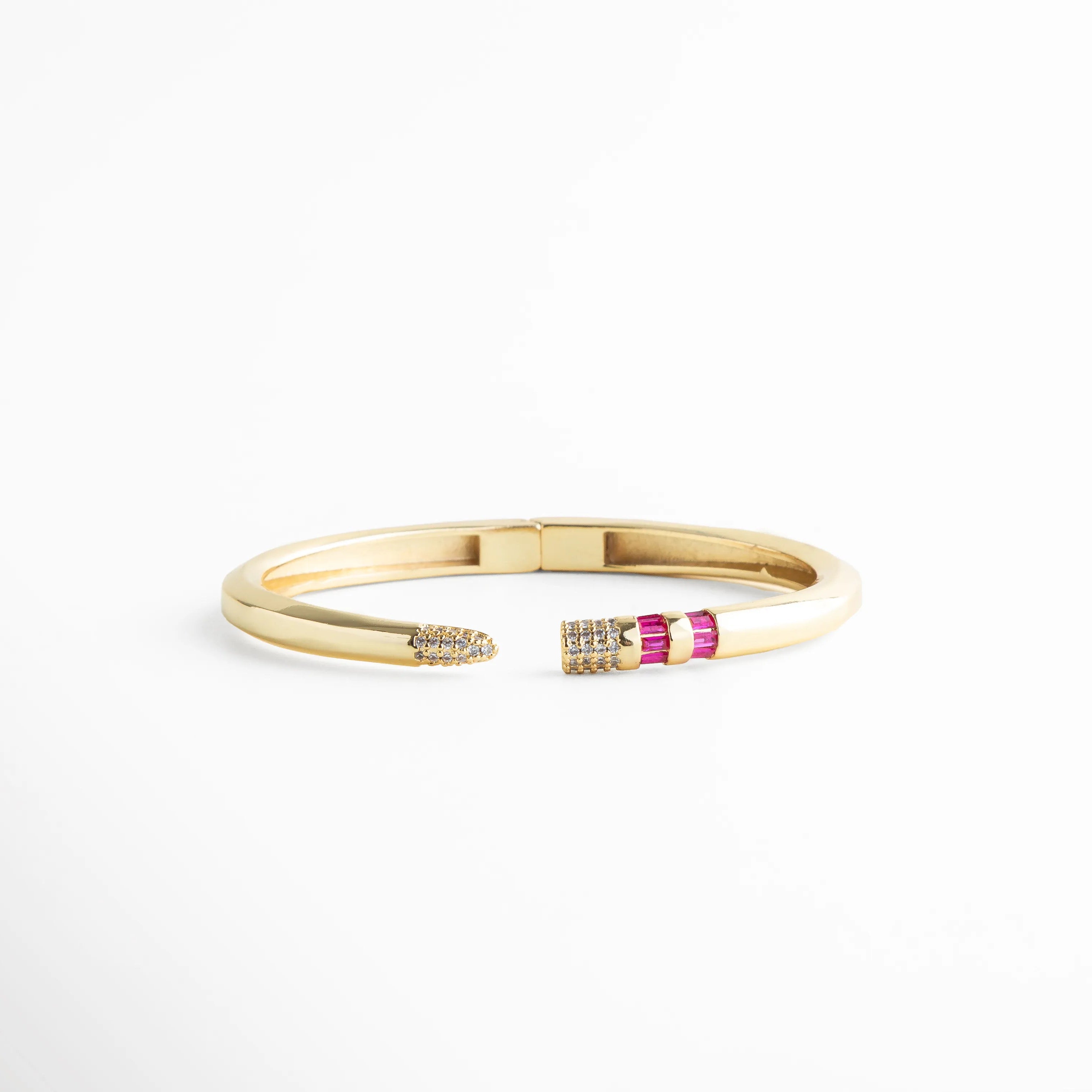Brazalete rígido dorado con puntas abiertas decoradas con circonias fucsia y blancas, Pulsera de cobre con chapa de oro 18K y detalle de piedras baguette en color magenta, Vista frontal de brazalete abierto con incrustaciones de circonia cúbica y acabado pulido, Joyería elegante: brazalete dorado con diseño de puntas asimétricas y cristales brillantes, Brazalete tipo "cuff" con bisagra central y detalles de pedrería fina en tonos rosa y cristal.