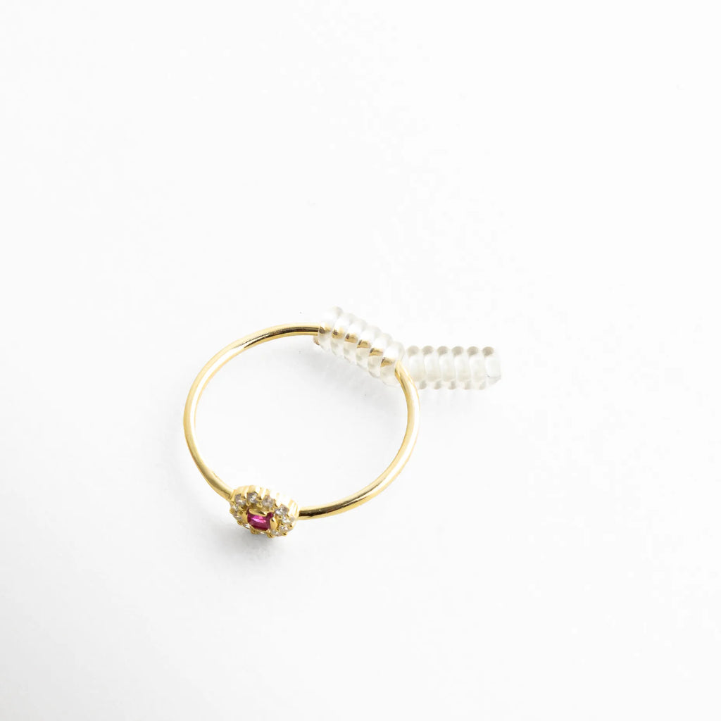 Anillo dorado con piedra central color rubí rodeada de cristales brillantes, acompañado de un discreto resorte transparente que ajusta su tamaño con elegancia. La imagen resalta el contraste entre el brillo del metal dorado y la sutileza del accesorio, ideal para mostrar cómo se adapta sin perder estilo.