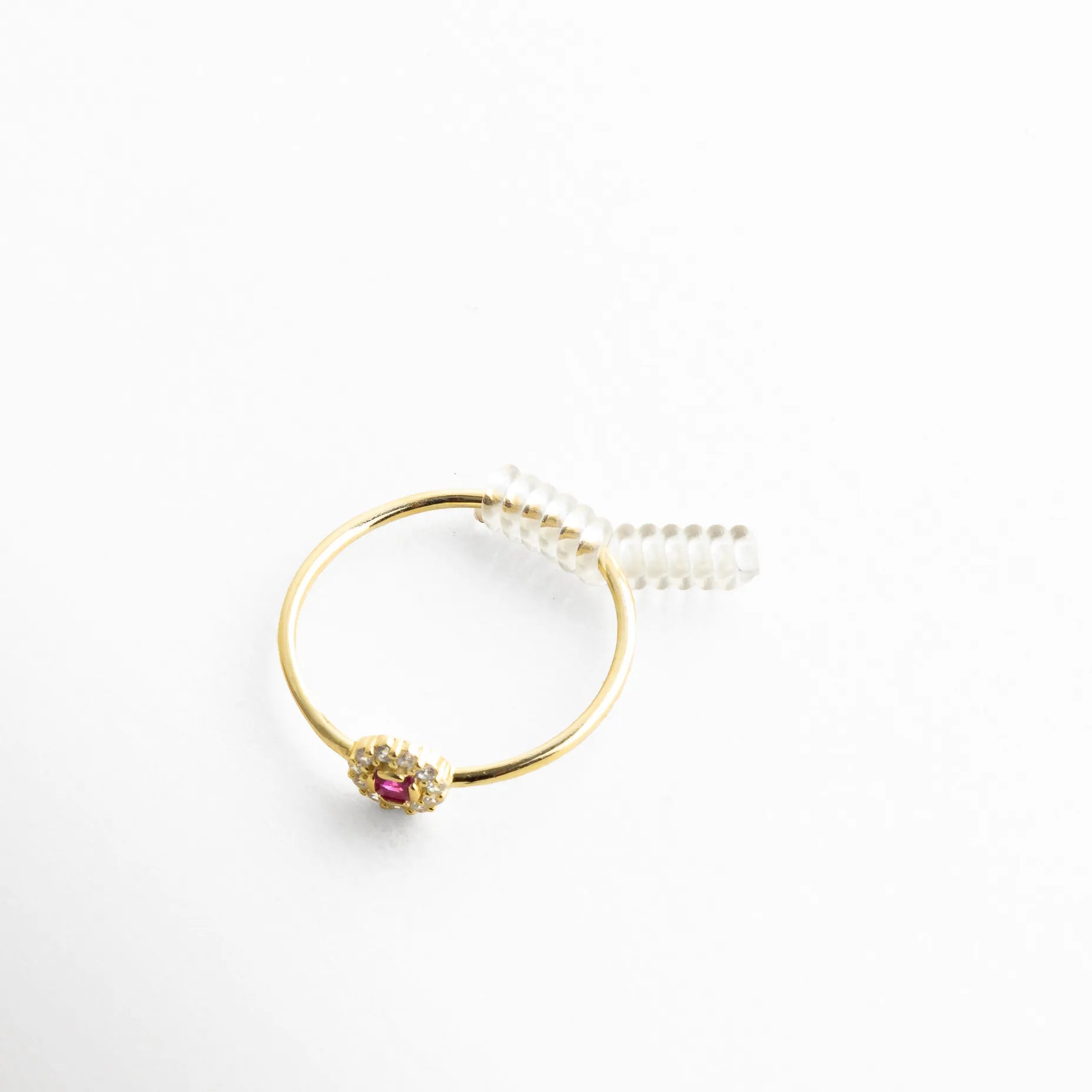 Anillo dorado con piedra central color rubí rodeada de cristales brillantes, acompañado de un discreto resorte transparente que ajusta su tamaño con elegancia. La imagen resalta el contraste entre el brillo del metal dorado y la sutileza del accesorio, ideal para mostrar cómo se adapta sin perder estilo.