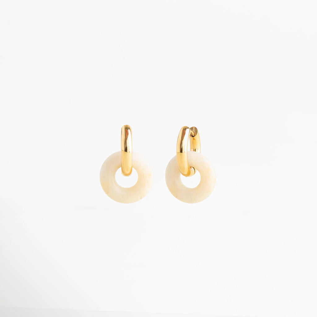 Par de arracadas Emilia Hoops mostrando la simetría de la piedra natural y el herraje dorado, Vista de catálogo de aretes dorados con dijes circulares de piedra natural de 2.0 cm de diámetro, Pendientes de acero inoxidable enchapados en oro con piedras naturales en tonos neutros, Diseño minimalista de aretes Hoops con colgantes de piedra orgánica para un look "effortless", Perspectiva frontal de los Emilia Hoops resaltando la proporción del herraje y la piedra.