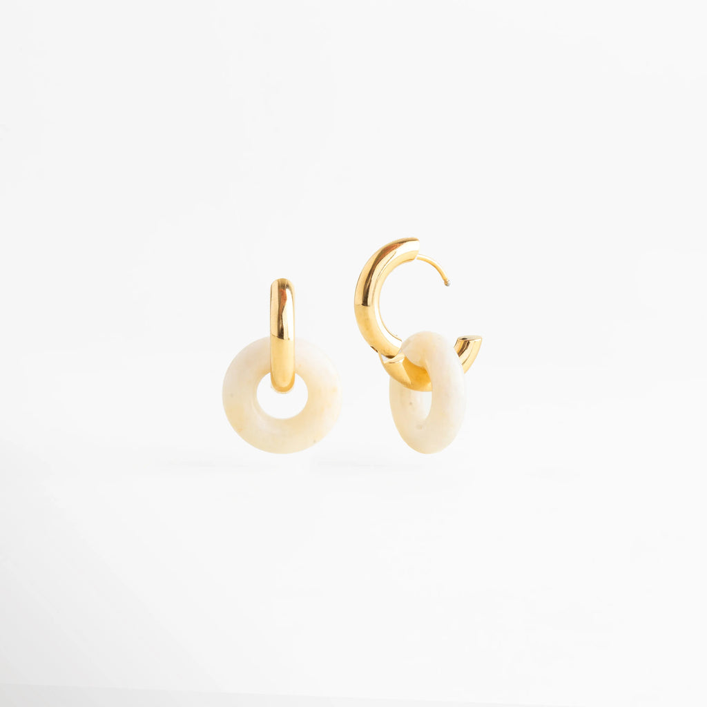 Vista lateral de los aretes Emilia Hoops mostrando el sistema de cierre de bisagra dorado, Pendientes de acero inoxidable con baño de oro abiertos para mostrar la funcionalidad del broche, Detalle del perfil del dije de piedra natural y el grosor del aro dorado de 0.4 cm, Perspectiva angular de las arracadas con piedra natural resaltando el acabado pulido espejo, Fotografía de producto mostrando el mecanismo de seguridad de los aretes dorados Emilia.