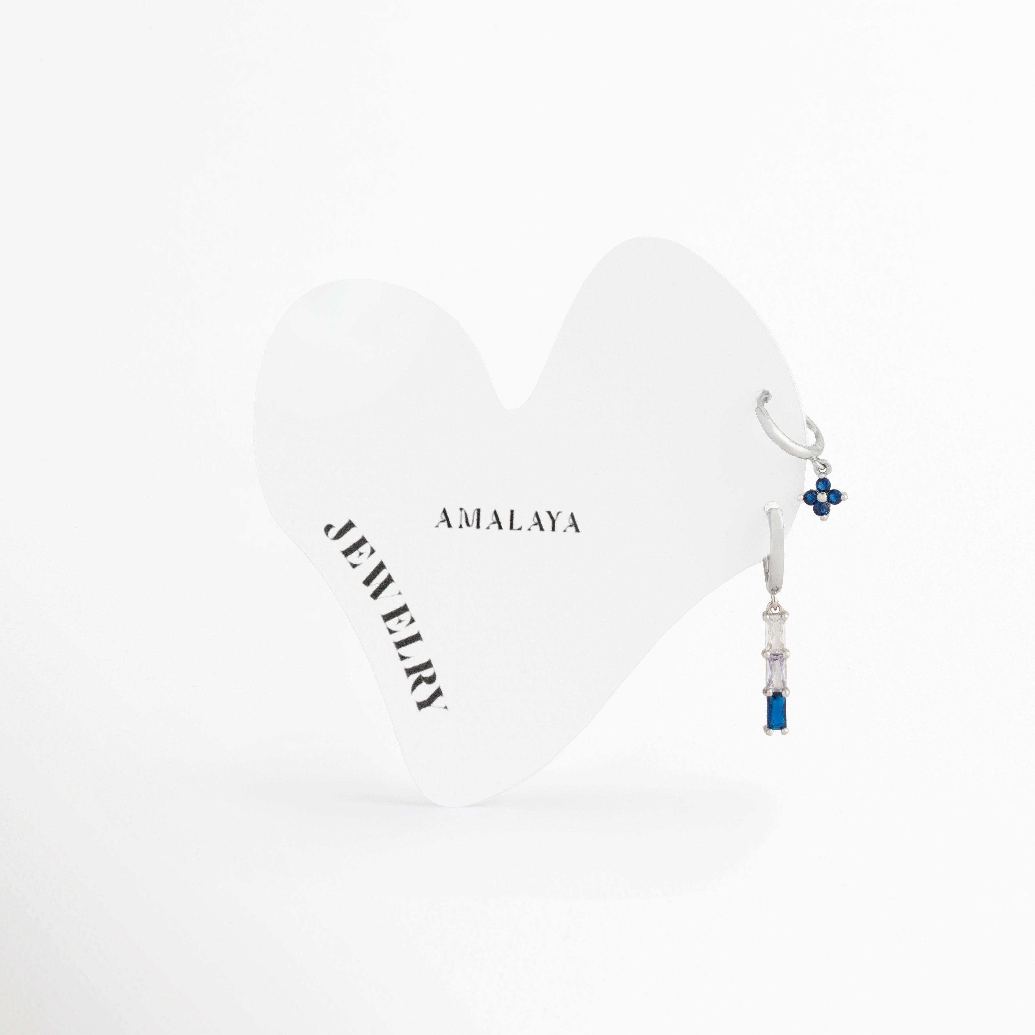 Dúo de aretes dorados en oro blanco de acero inoxidable con flor azul y barra colgante en degradado, Dos huggies plateados con zirconias de color azul y lavanda sobre tarjeta blanca de Amalaya Jewelry, Joyería de acero inoxidable hipoalergénica: mini arracadas con dijes colgantes de flores y piedras baguette, Aretes resistentes al agua color plata con piedras de colores para múltiples perforaciones, Detalle de combinación de aretes asimétricos con degradado púrpura y azul en oro blanco sobre fondo blanc